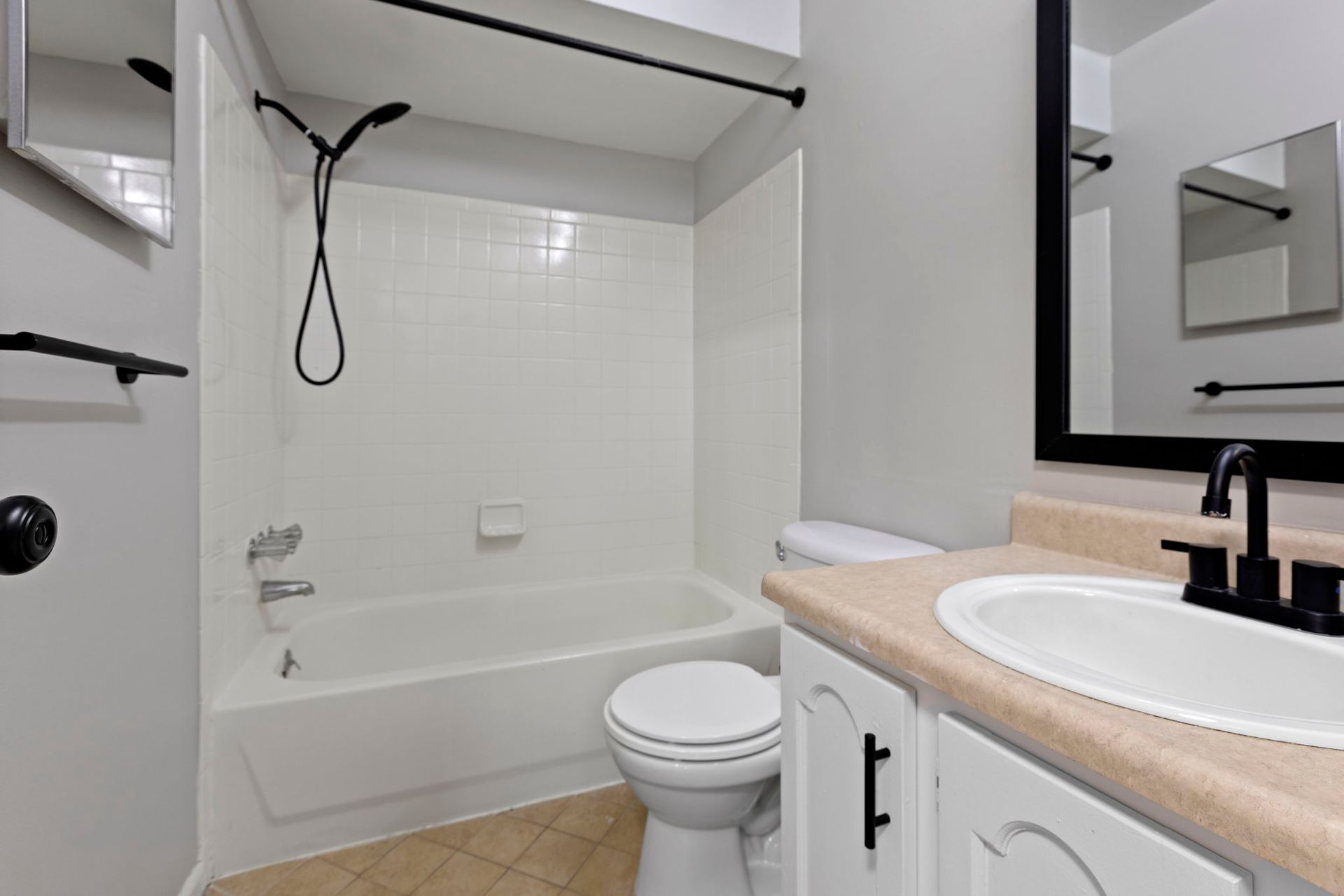 Small bathroom with white tub and toilet, beige counter, and black fixtures.