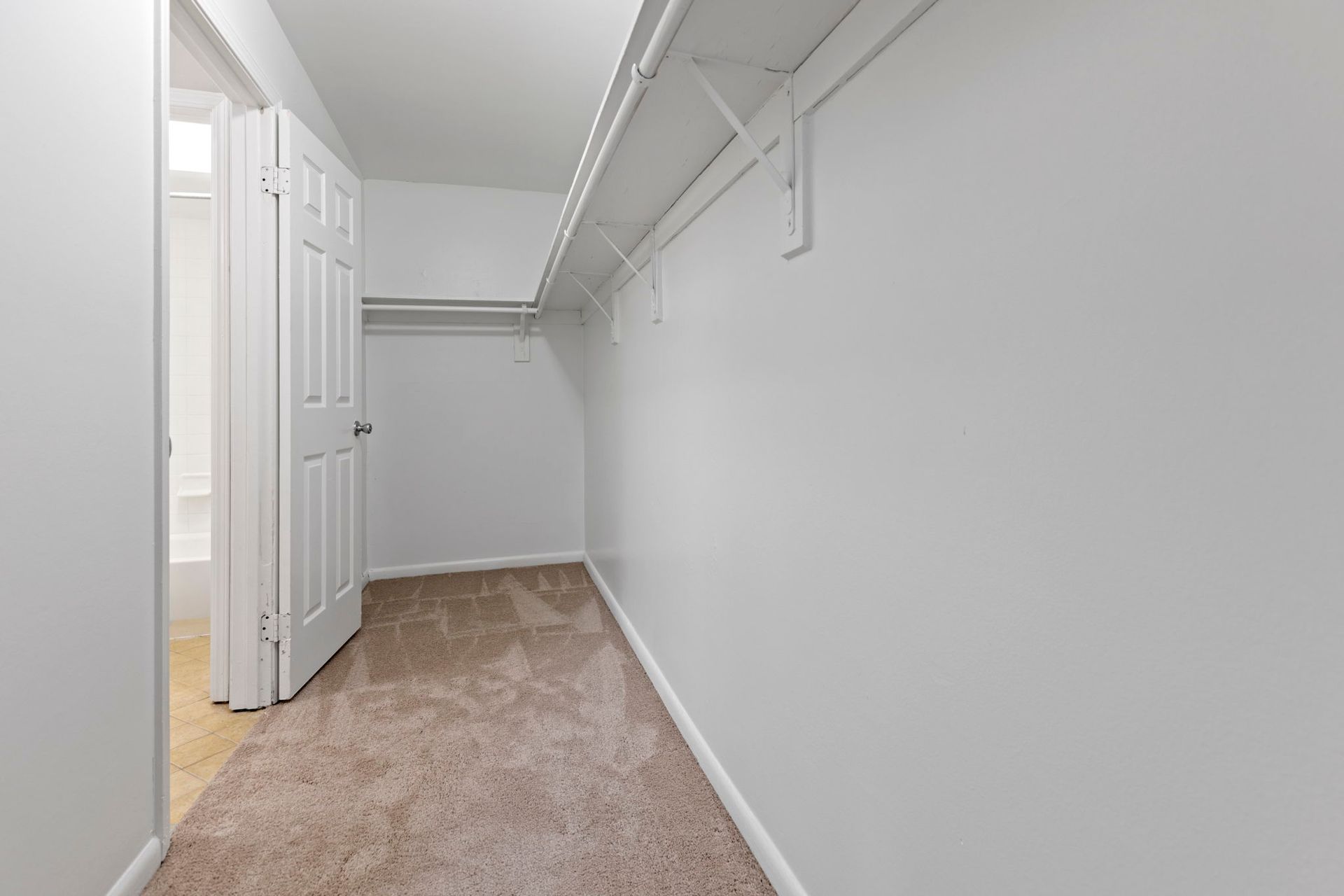 Empty walk-in closet with white walls, carpet, and a door leading to a bathroom.