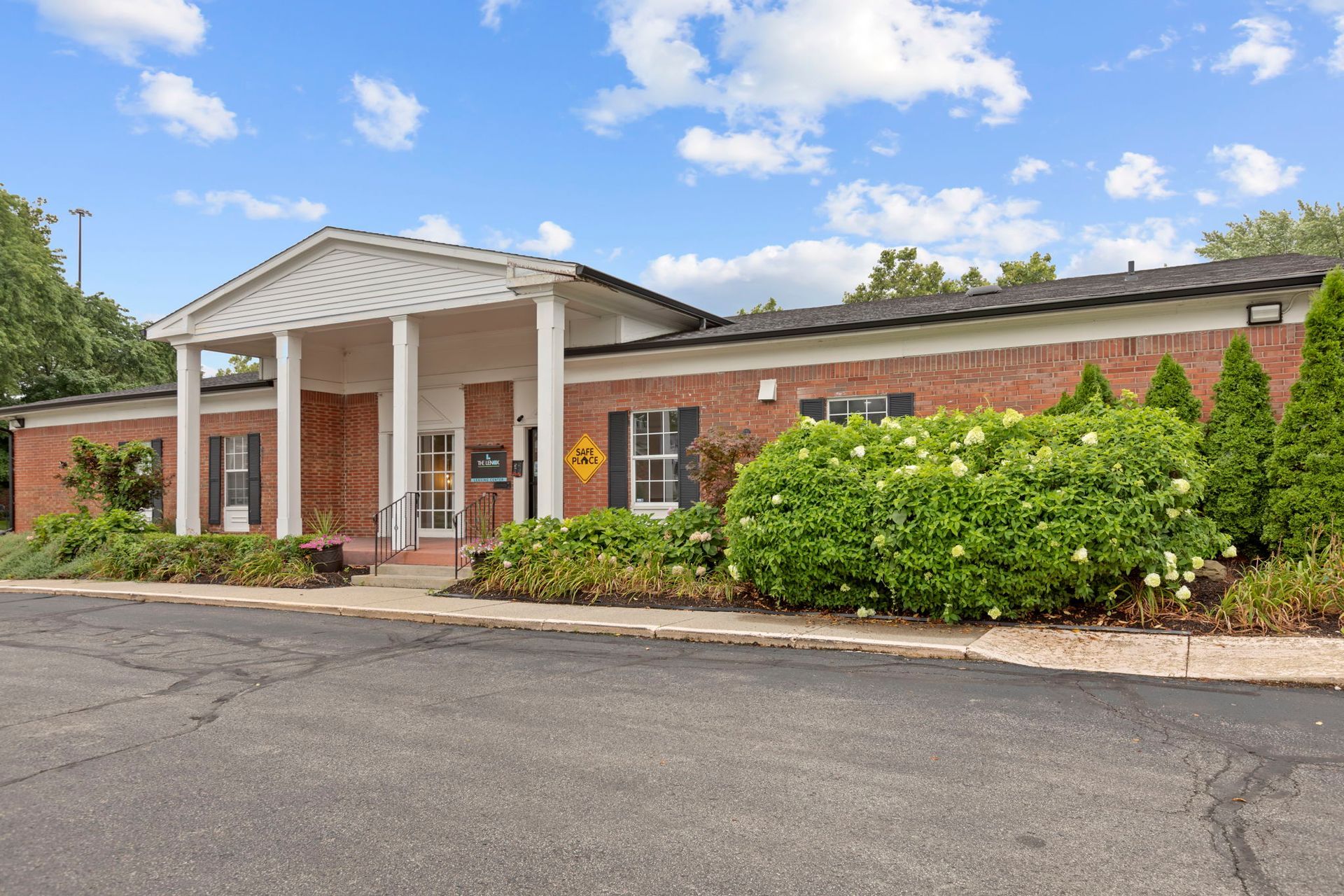 Brick building with white columns and a peaked roof. Green bushes and blue sky visible.
