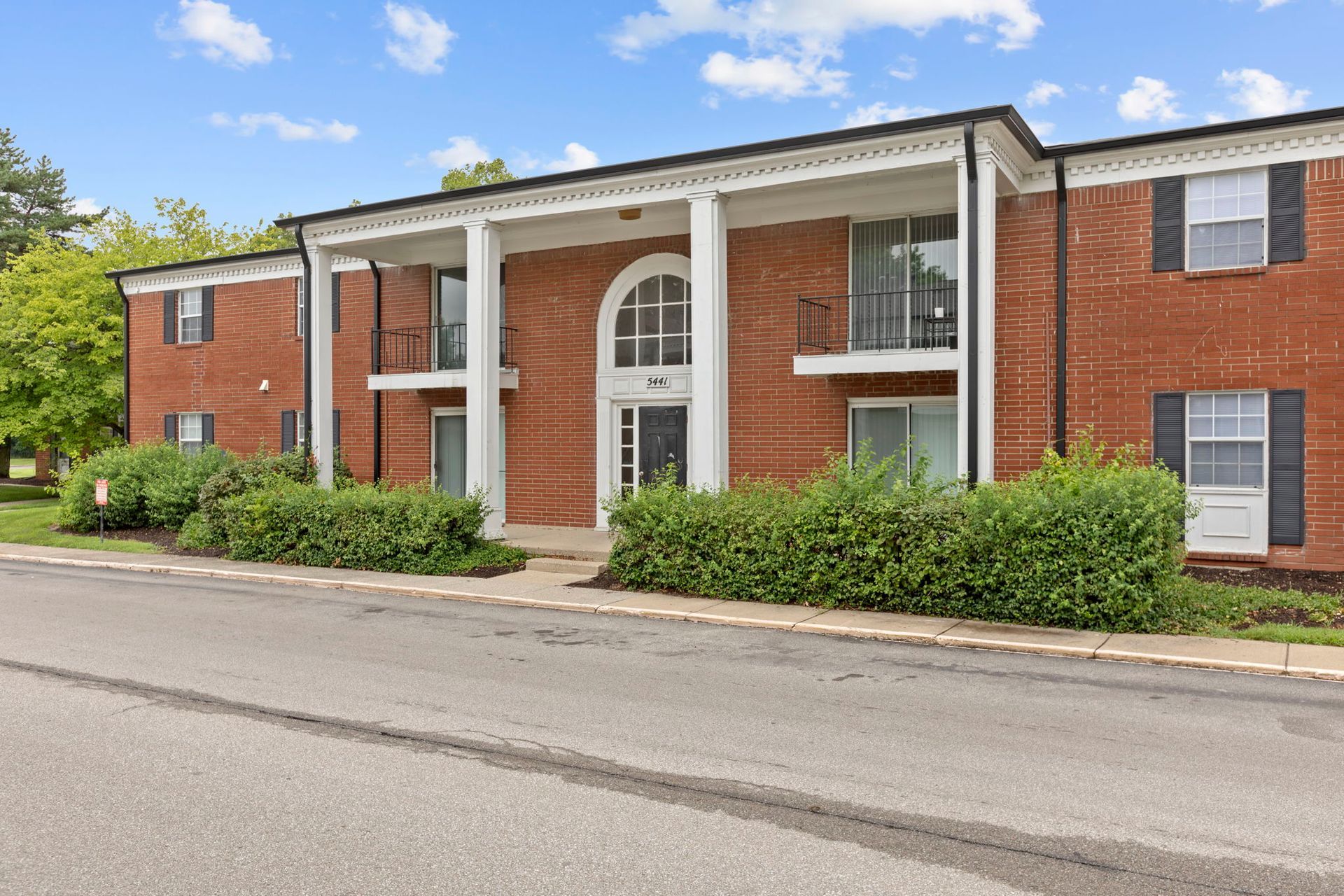 Red brick apartment building with white columns and a central arched doorway; a paved road in the foreground.