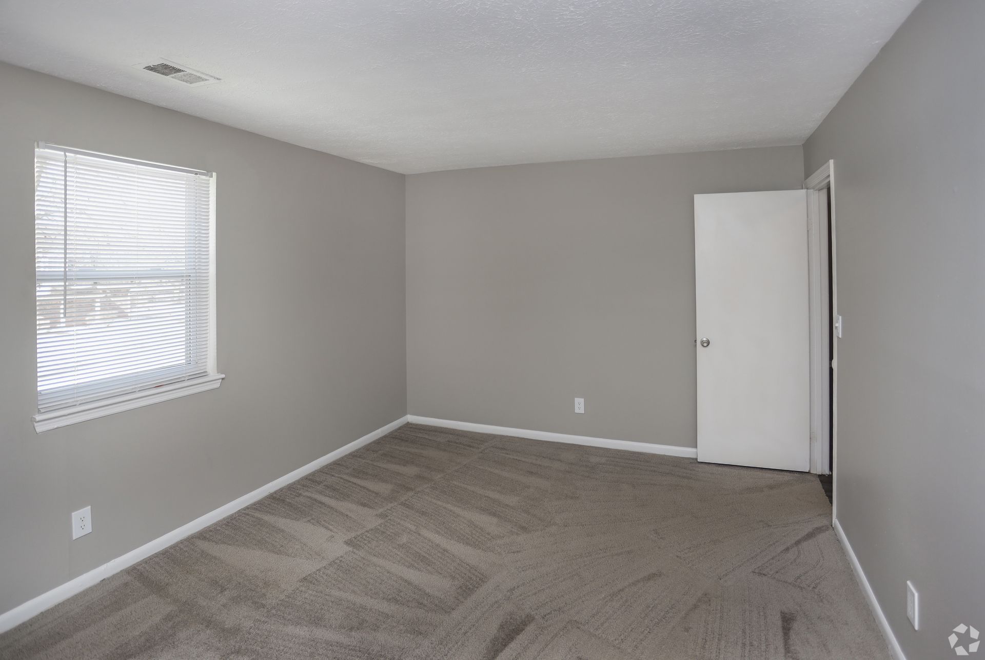 Empty room with gray walls, carpet, and white trim. Window with blinds and a closed door.