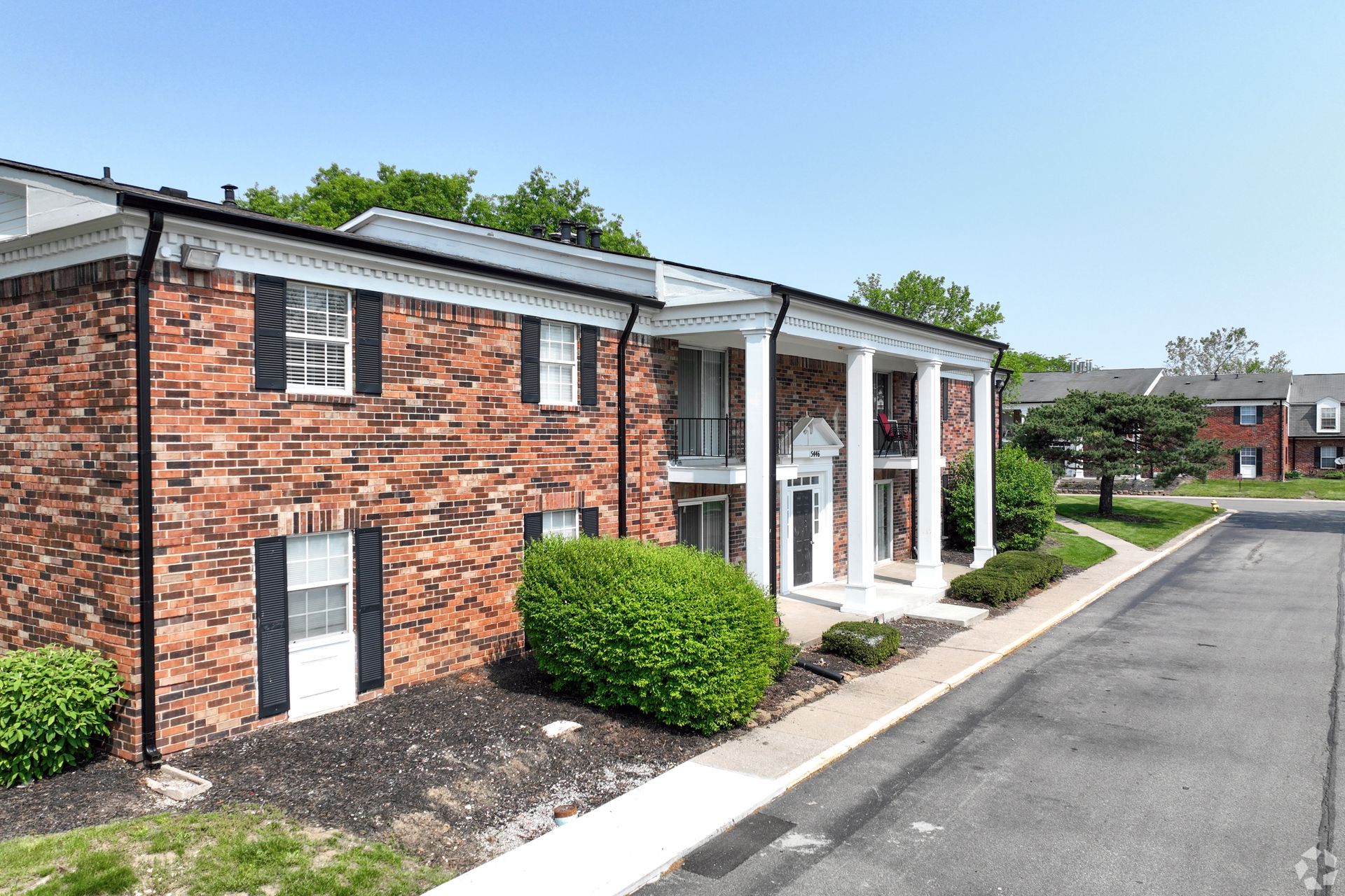 Brick apartment building with white columns, black shutters, and landscaping along a paved road.