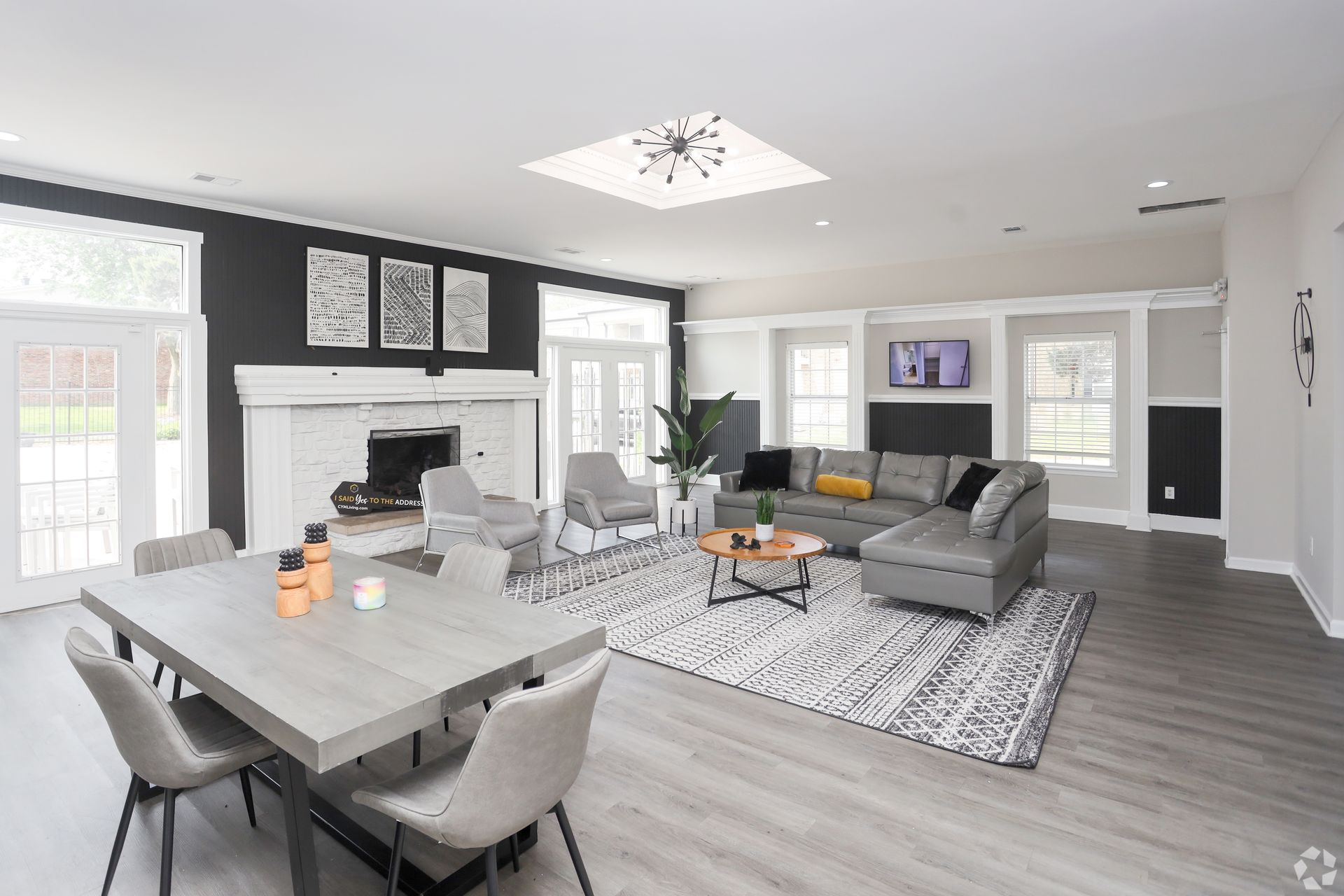 Spacious, modern living room with dining area. Gray sofa, rug, and table. White fireplace and walls; black accent wall.