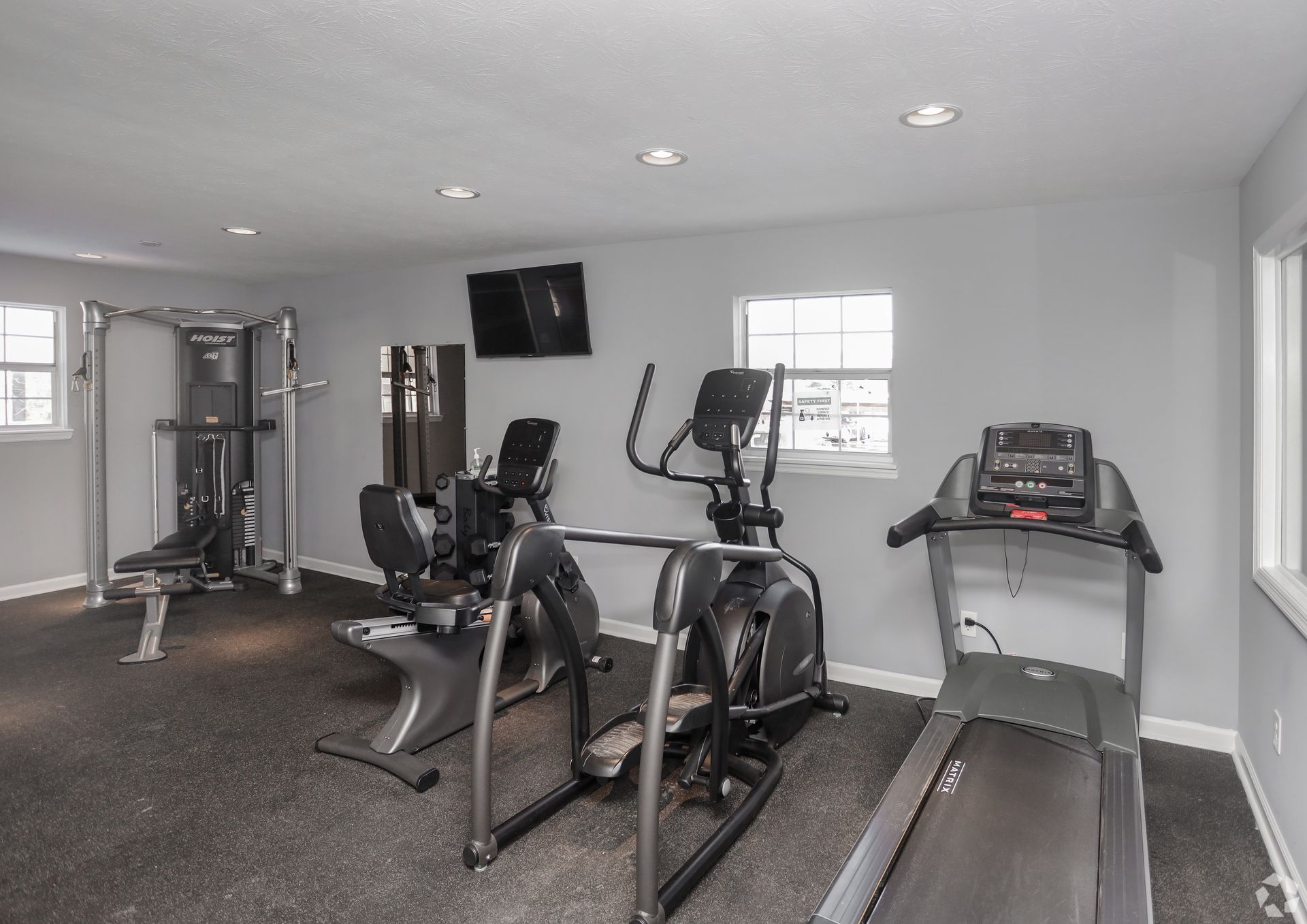 Gym with workout equipment, including a treadmill, elliptical, and weight machines. Gray walls and black flooring.