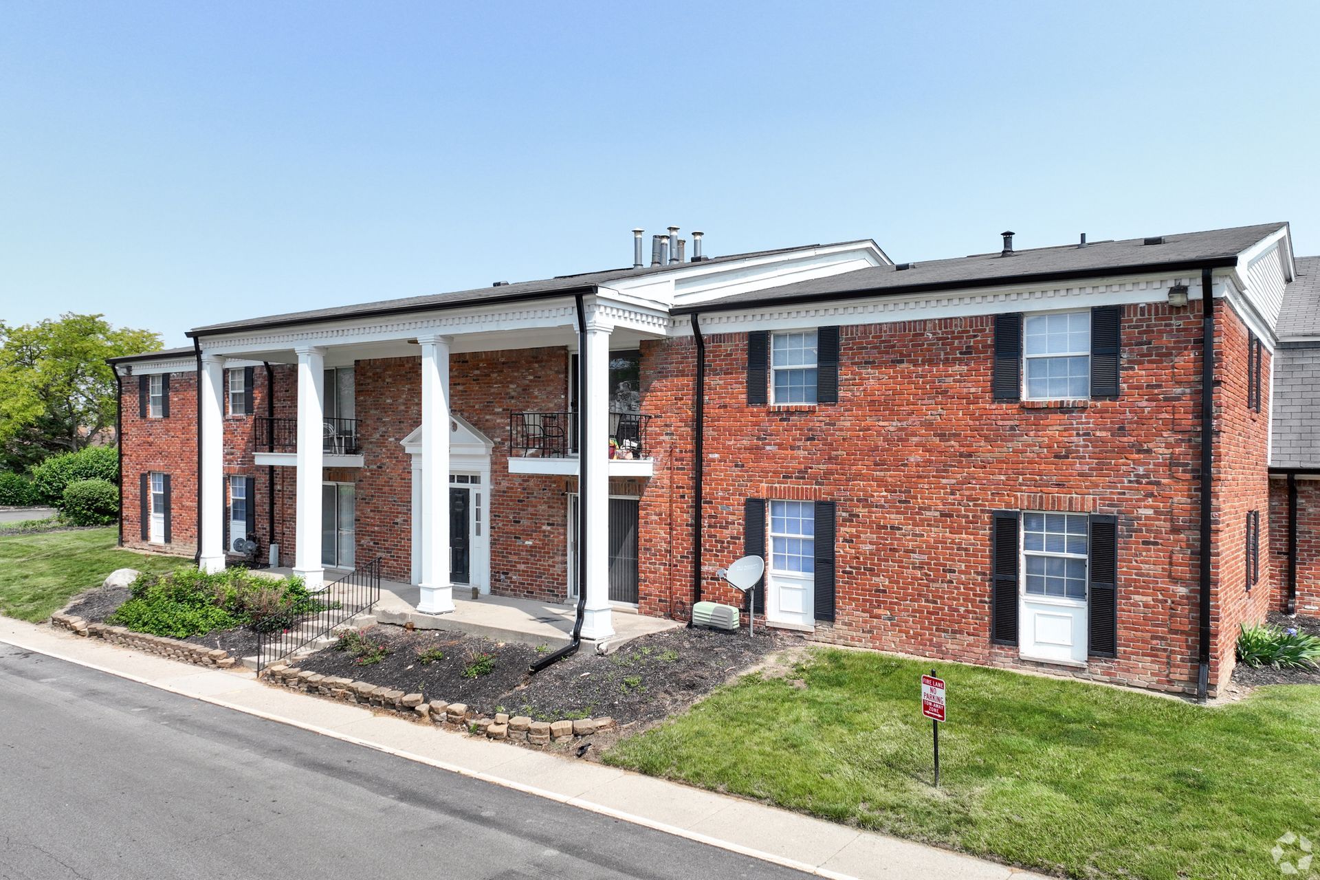Red brick apartment building with white columns and black shutters under a blue sky.