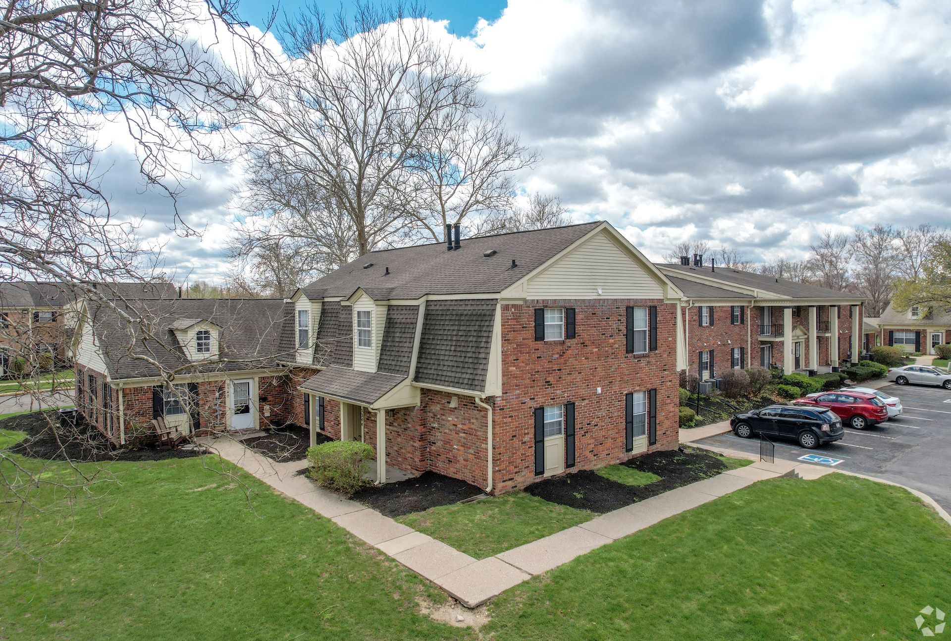 Apartment buildings with brick exteriors, green lawn, and parking area on a cloudy day.