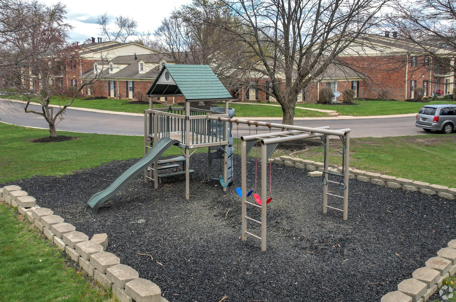 Playground with slide and monkey bars on black rubber mulch, in front of apartment buildings.