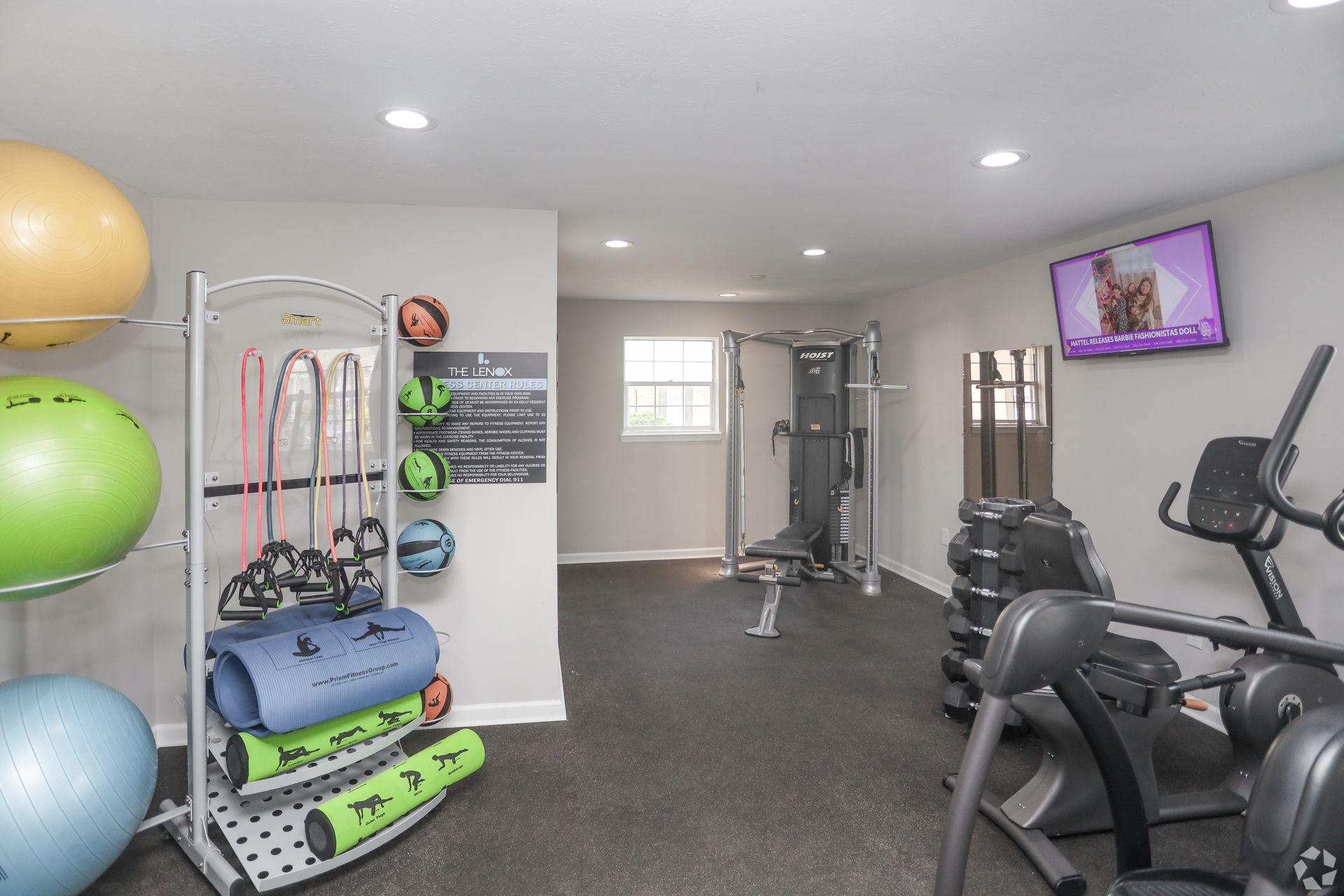 Gym with exercise equipment: balls, weights, resistance bands, and a bench press. A TV is on the wall.