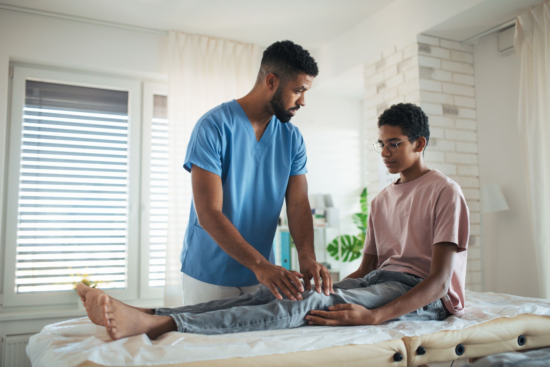 Healthcare provider examining patient's knee in light room with window. Both are dressed in casual clothing.