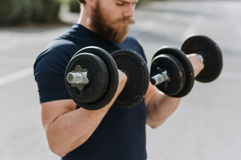 A bearded man is lifting two dumbbells on his biceps.