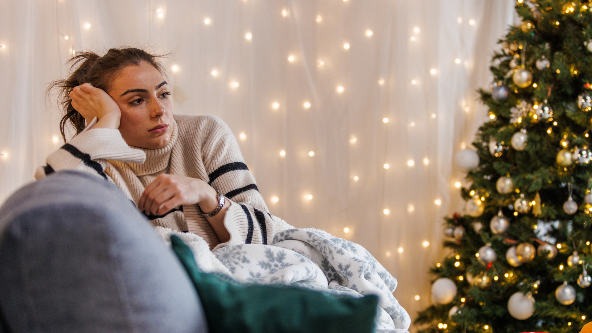 woman sitting on couch next to Christmas tree looking sad