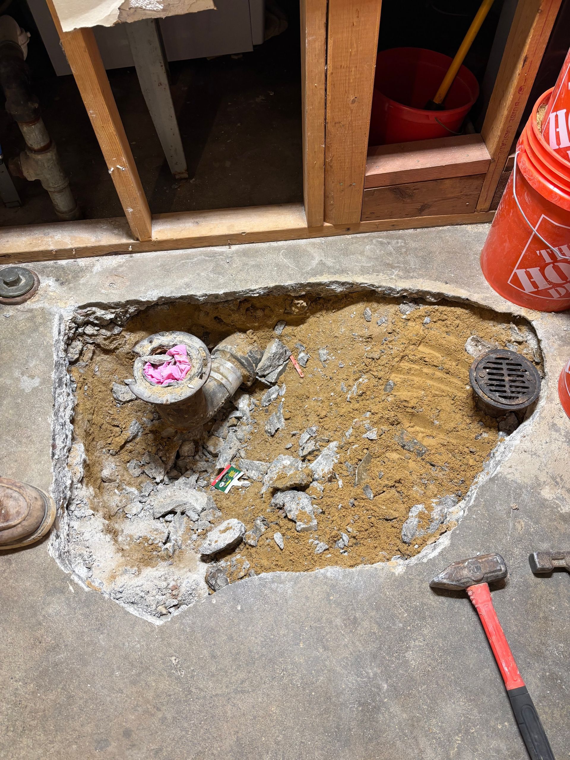 A hole in the floor next to a red bucket and a hammer.