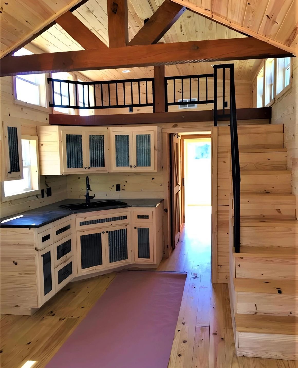 Tiny house interior with kitchen, loft, and stairs. Light wood, black accents, pink rug, open doorway.