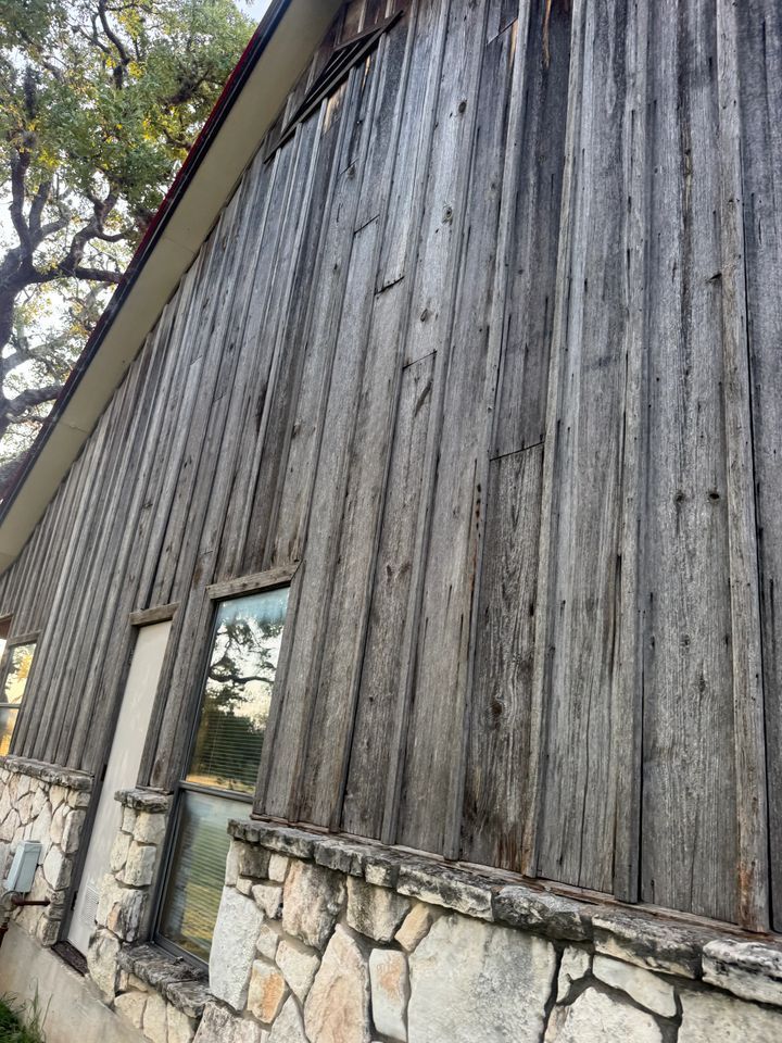 Weathered wood siding on a building with a stone base, a small window, and a view of trees.