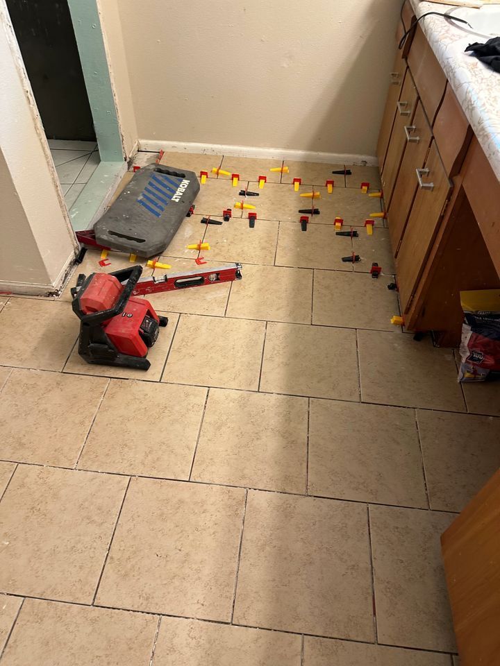 Bathroom floor tiling in progress: tan tiles with spacers and tools, near cabinets and a doorway.