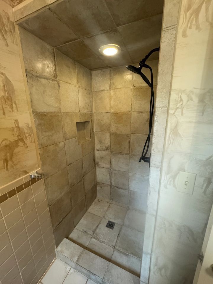 Small shower with beige tile and a black showerhead. Beige walls with a mural.