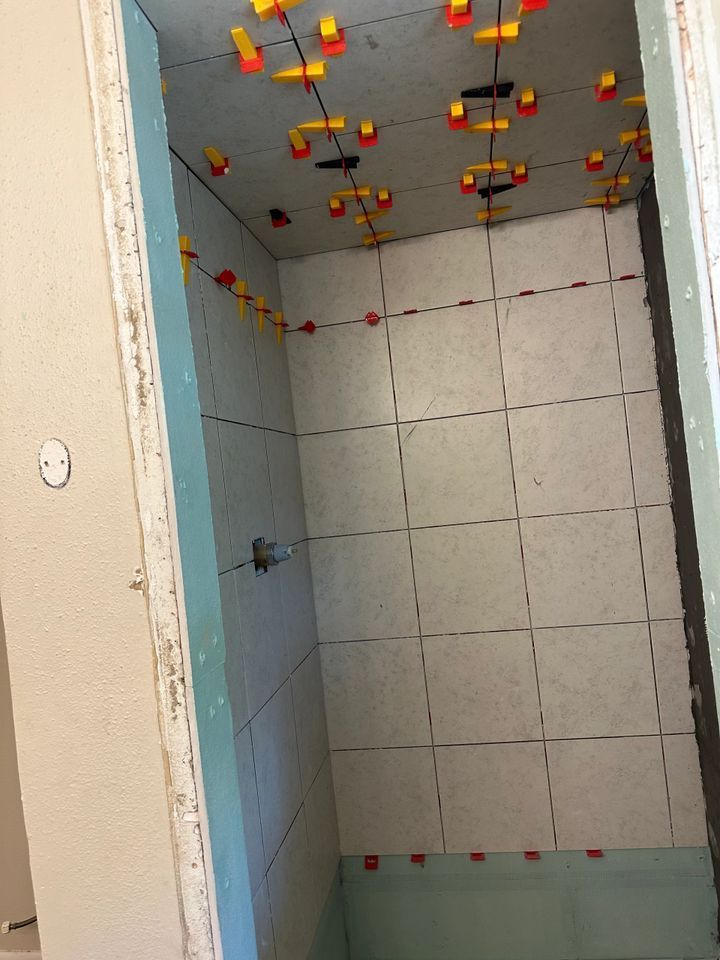 Tiled shower stall, with grout lines and leveling clips visible; green drywall and white tiles.