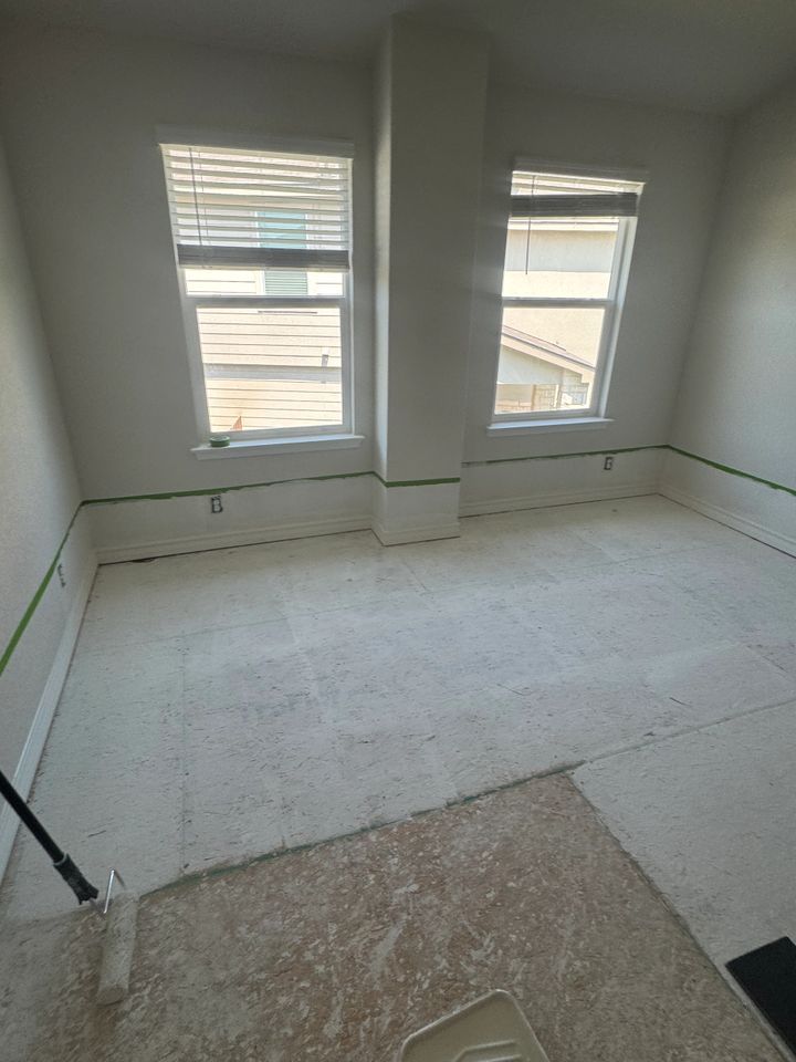 Room with two windows, white walls, floor partially covered with debris, and painter's tape.