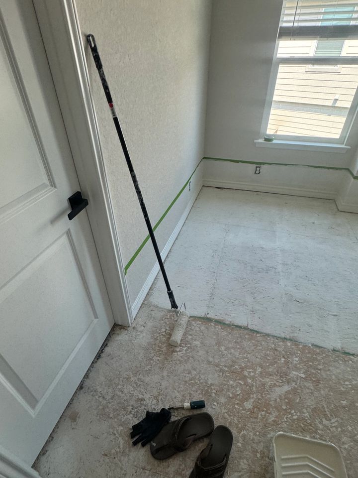 A room being painted: roller and handle propped, tape lines, floor covered in paint splatters, sandals and gloves on floor.