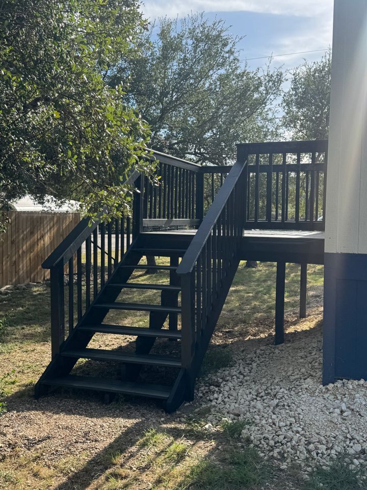 Black wooden deck with stairs, next to a building and gravel ground.