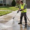 Person power washing a concrete driveway with a pressure washer, wearing safety gear outside a house.
