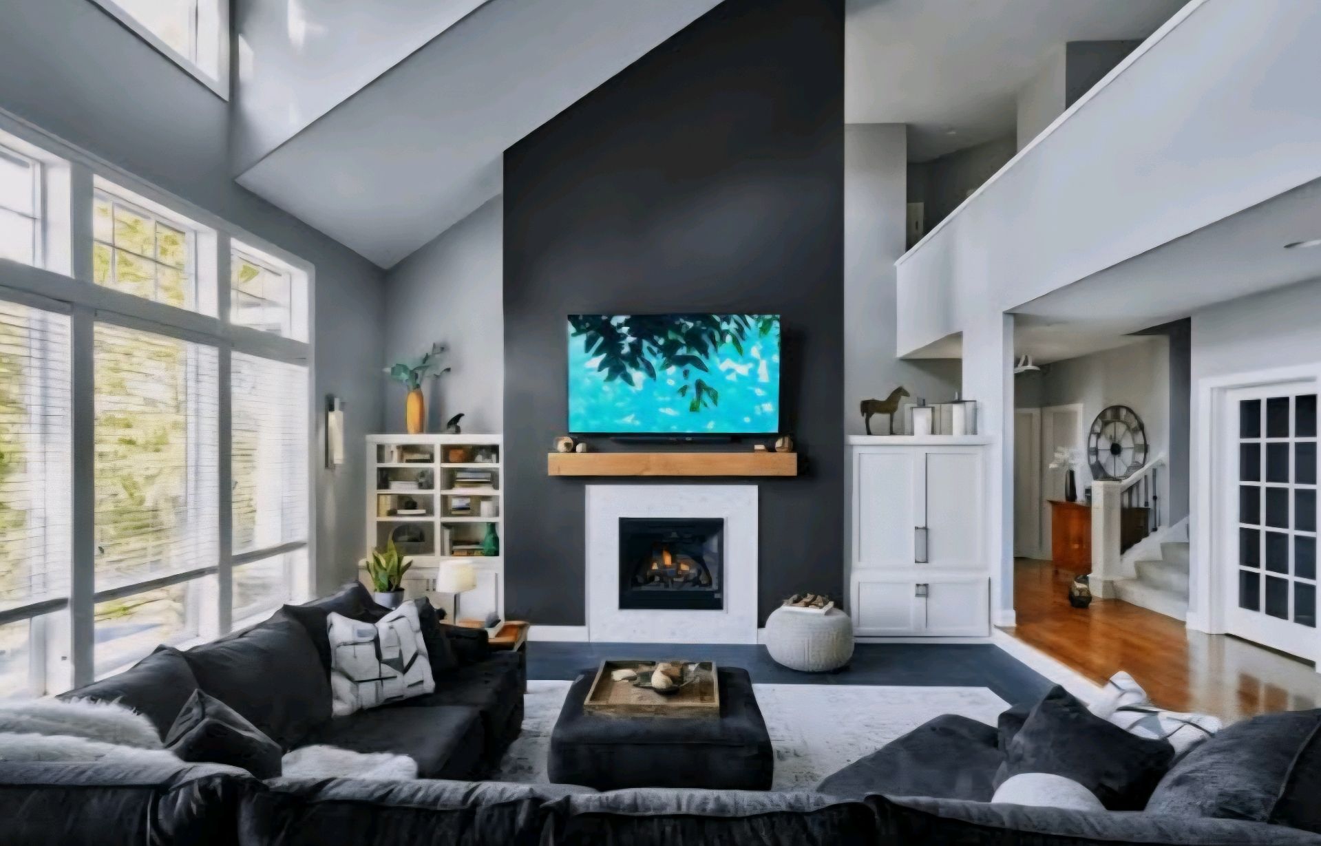 Living room with gray walls, dark sofa, large windows, and a fireplace with a mounted TV.