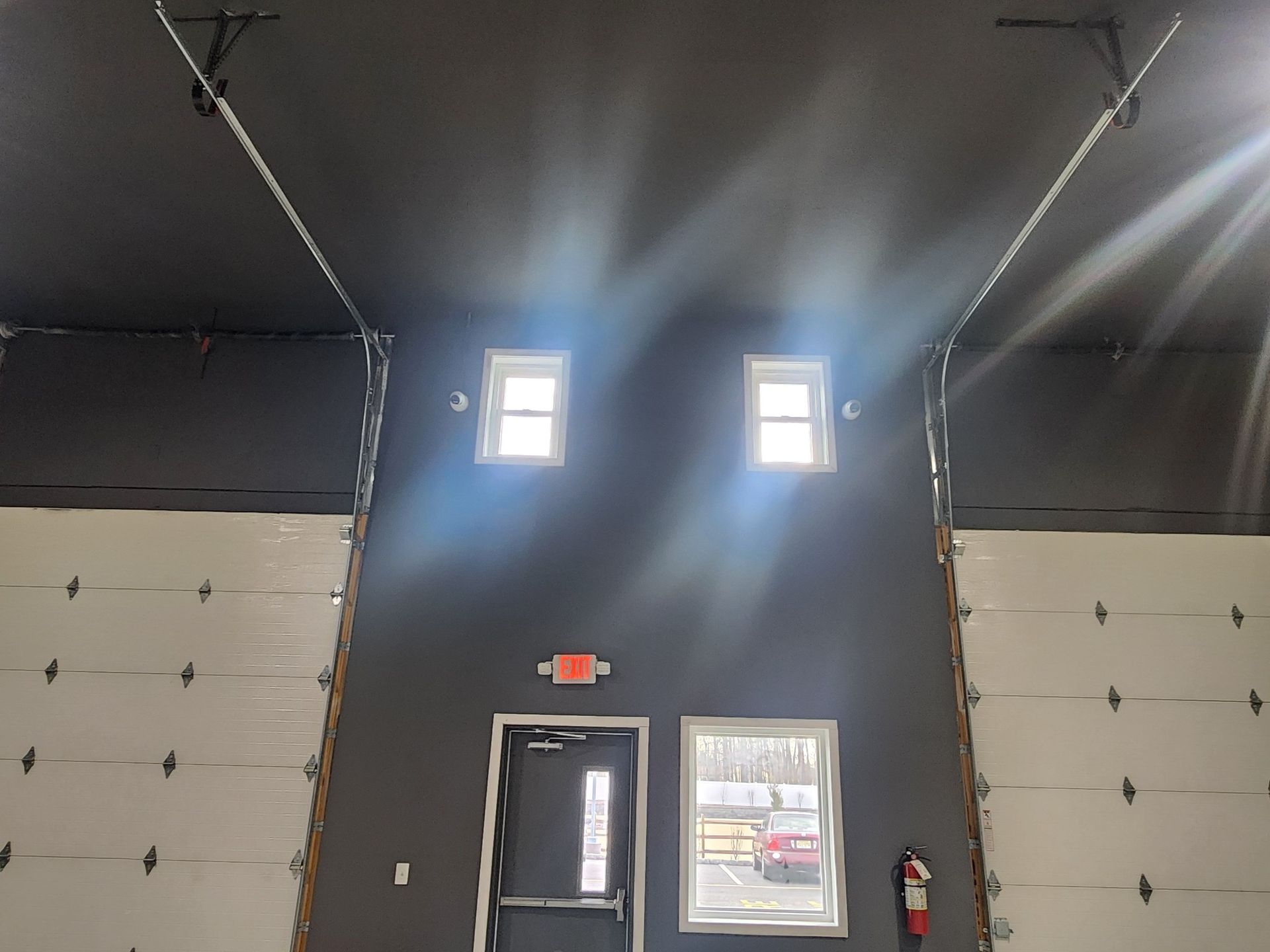 Interior view of a building with overhead garage doors and a central wall with windows, a door, and a fire extinguisher.
