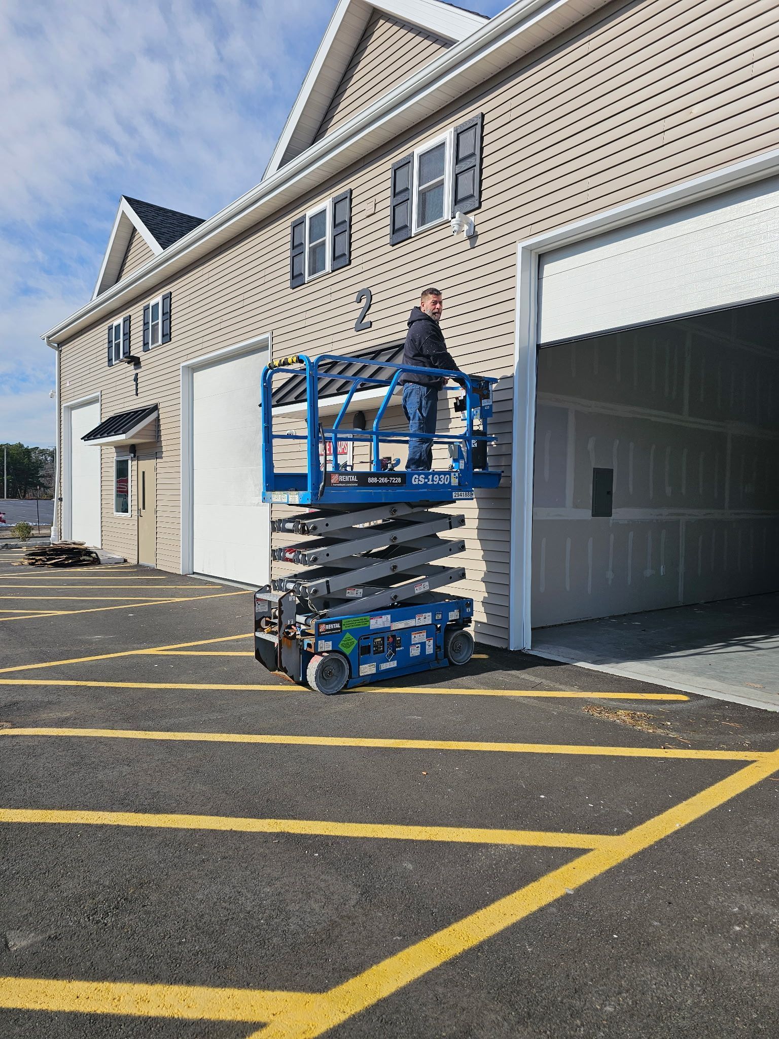 Person on a blue scissor lift near a building with an open garage door. Asphalt lot with yellow lines.