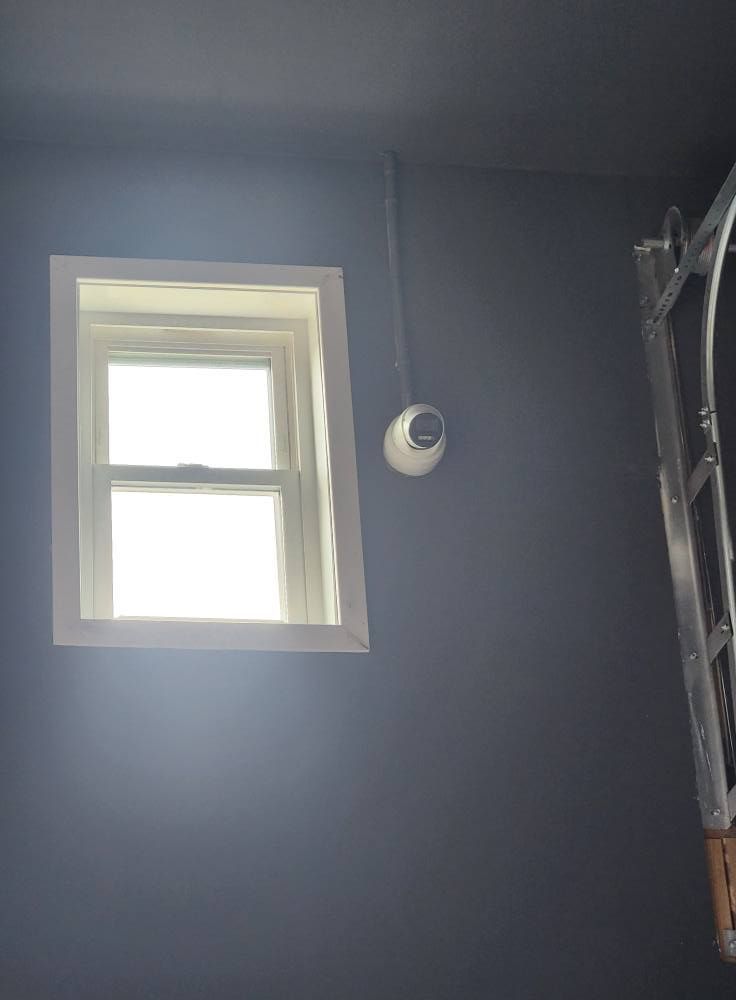 Window with white frame on gray wall, security camera below. Ladder partially visible on right.