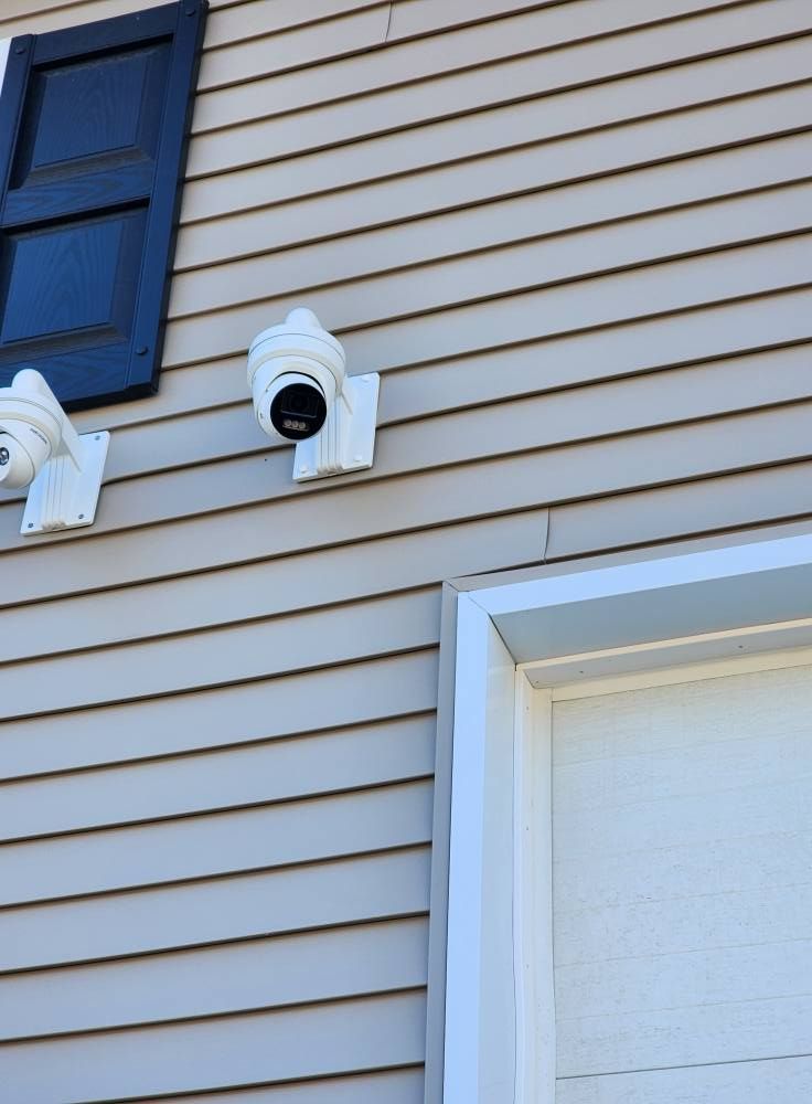 Two white security cameras mounted on beige siding next to a white door frame and a dark blue shutter.