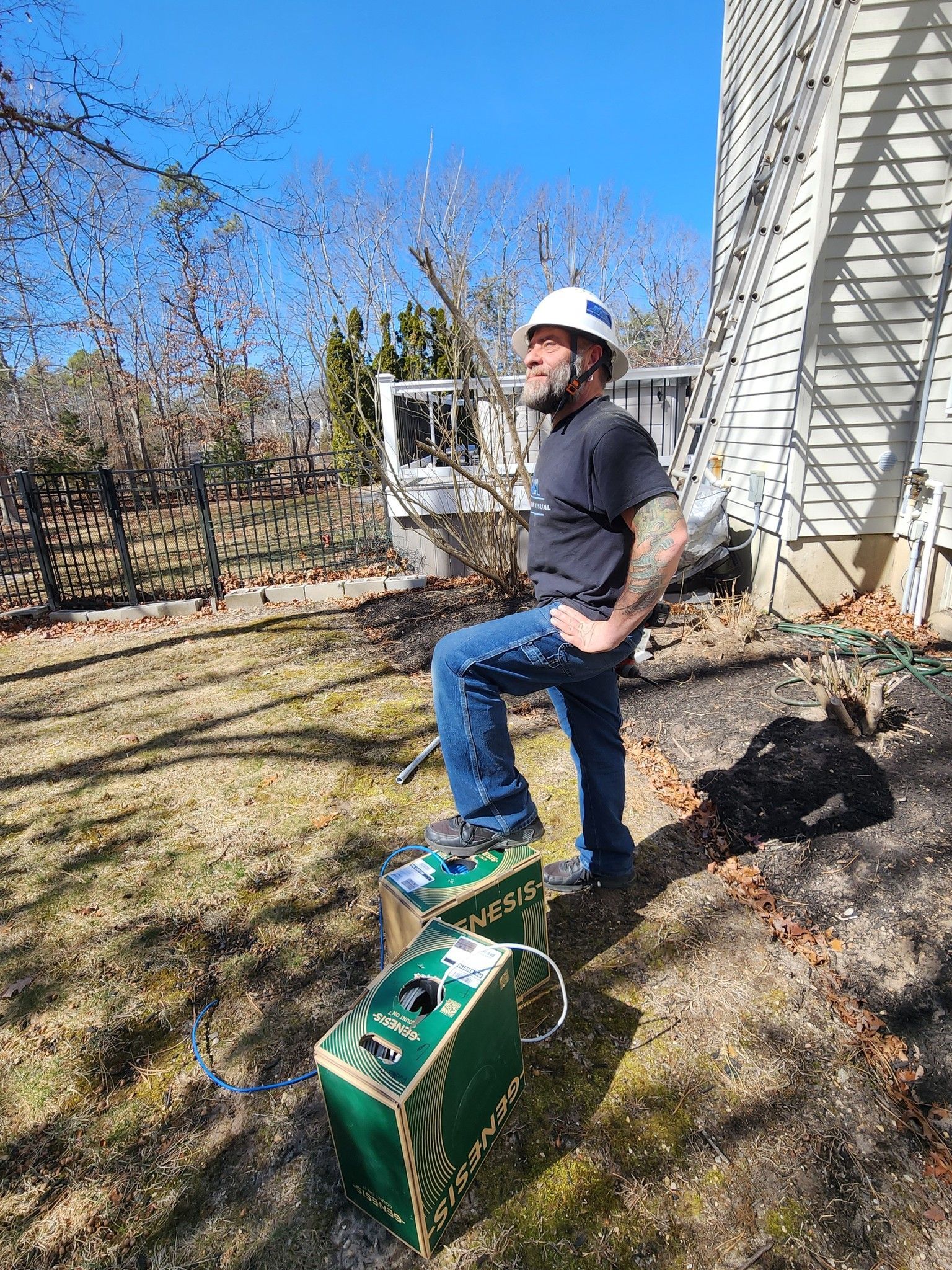 Man in hard hat stands on two green boxes outdoors near a house, sunny day.