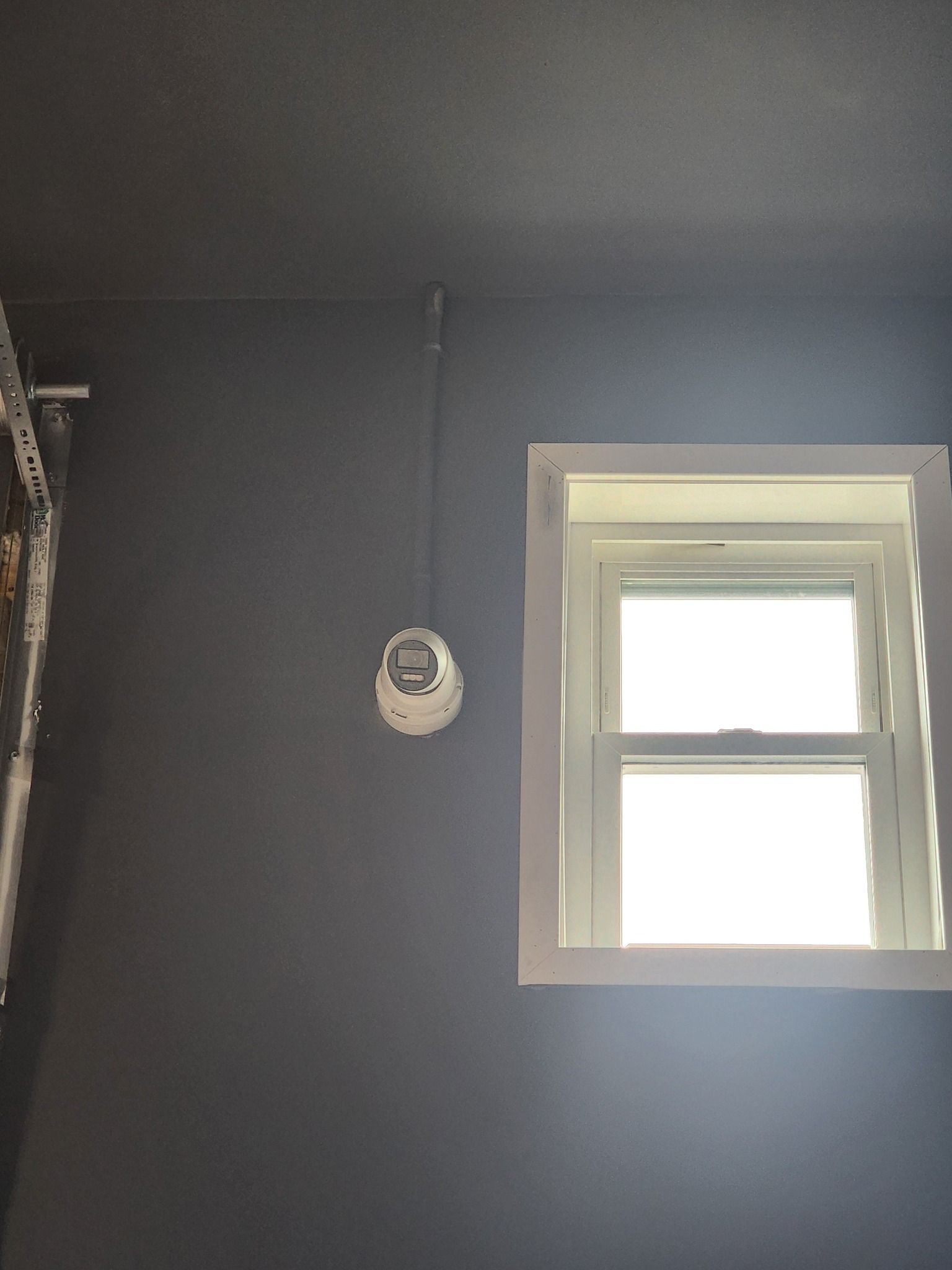White fire alarm on a dark gray wall, above a white-framed window.