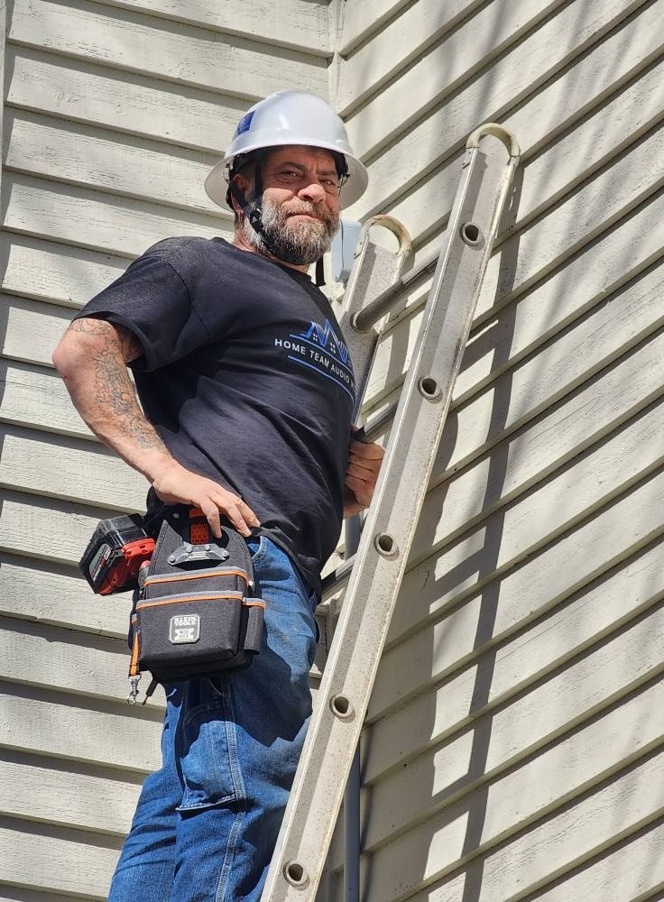 Man on a ladder wearing a hard hat, tool belt, and dark shirt working on a building exterior.