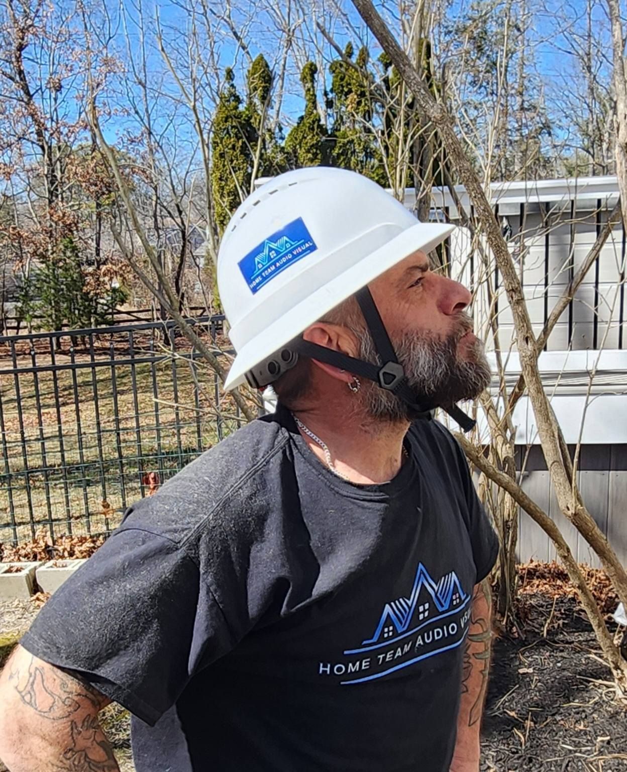 Man wearing a white hard hat looking upward. He's outdoors, near a fence and trees, wearing a black shirt.