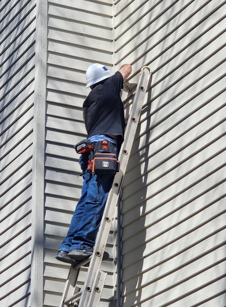 Person on a ladder, installing something on the siding of a building. They wear a hard hat and tool belt.
