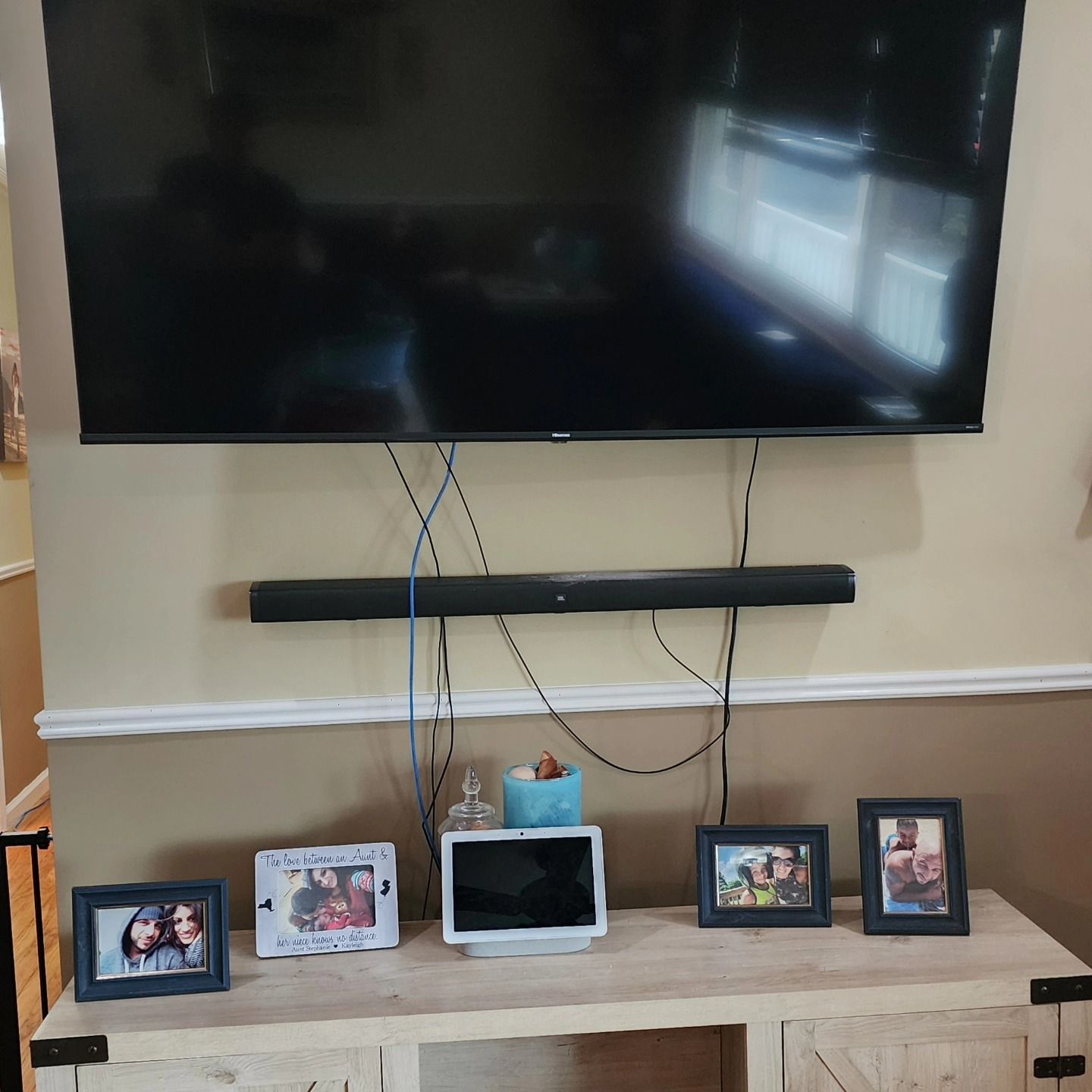 A TV and soundbar mounted above a light wood cabinet with framed photos and a smart display. Wires hang down.