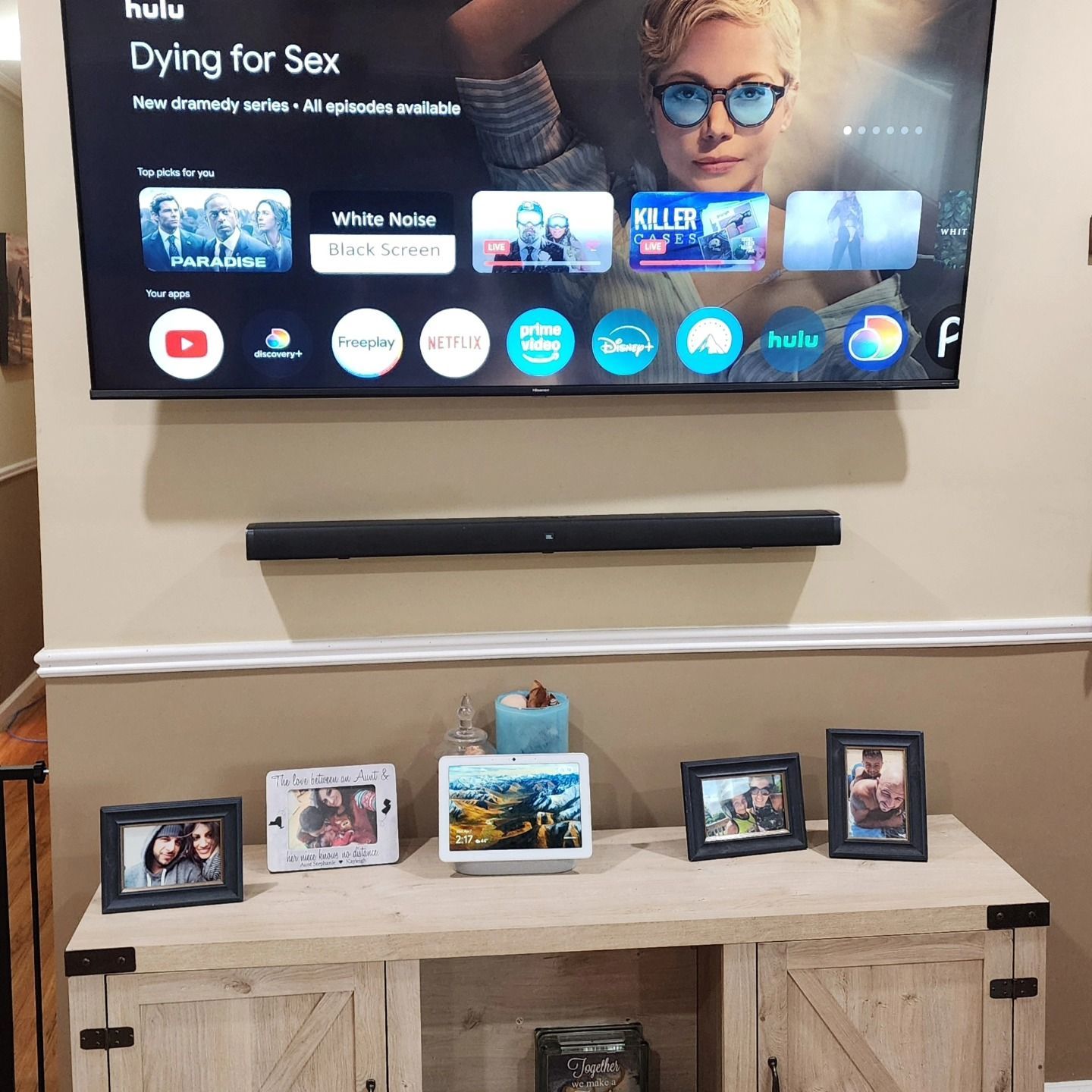 A TV above a soundbar, over a wooden cabinet with framed photos and a smart speaker.