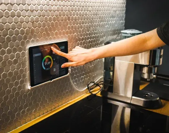 Person interacting with a tablet embedded in a tiled kitchen wall. Shows temperature control next to a coffee maker.
