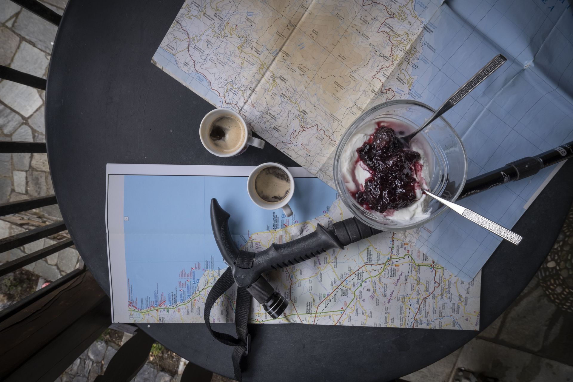 Coffee cups, map, yogurt with jam, ice axe on table, overhead shot.