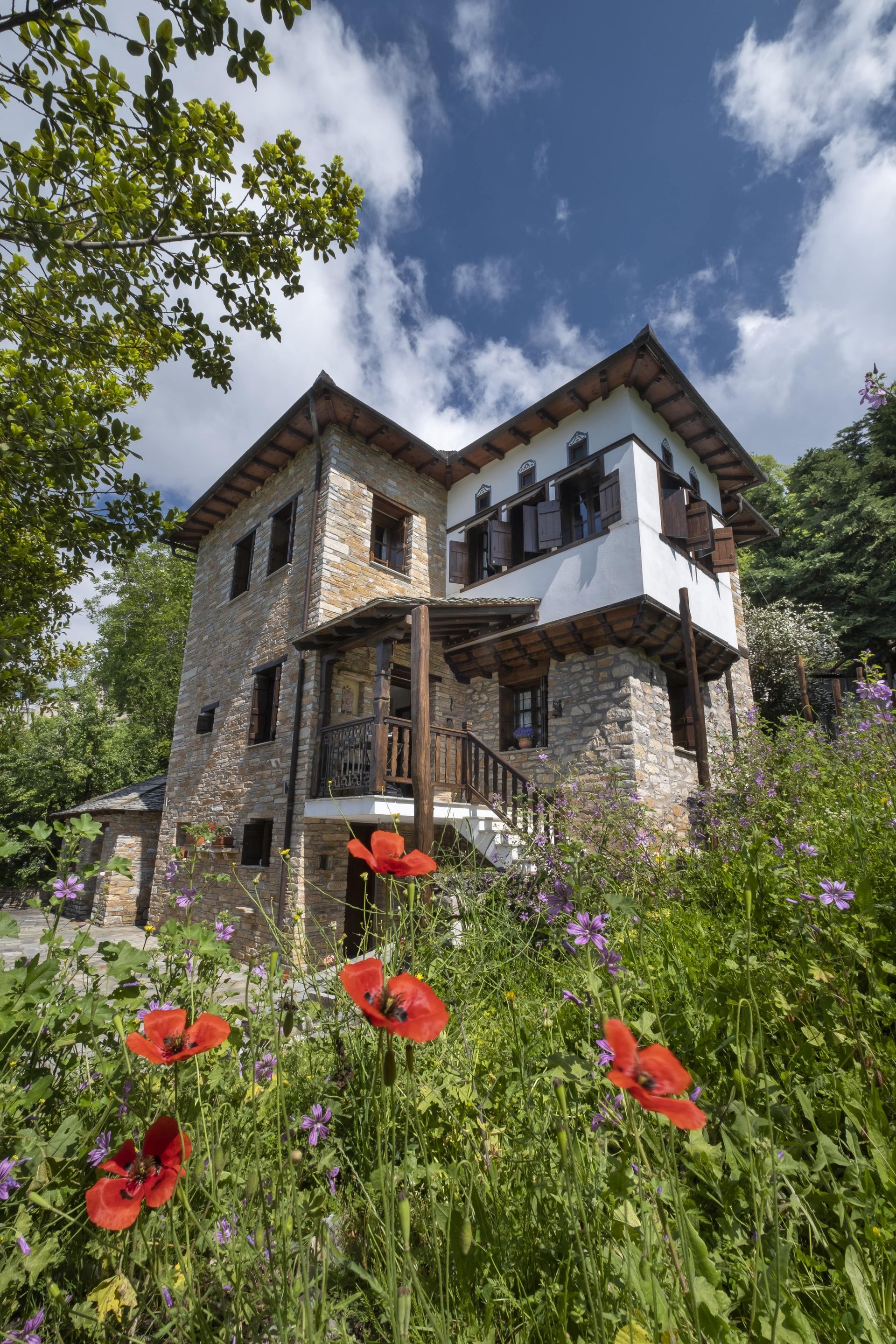 Stone house with red poppies, surrounded by greenery and blue sky with clouds.