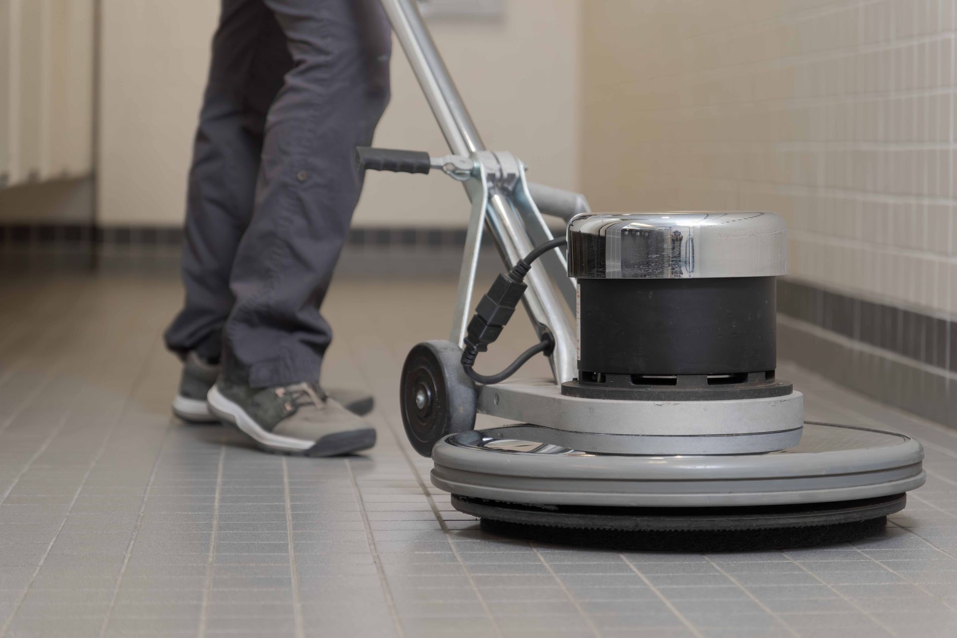 Person cleaning a tiled floor with a floor buffer in a tiled room.