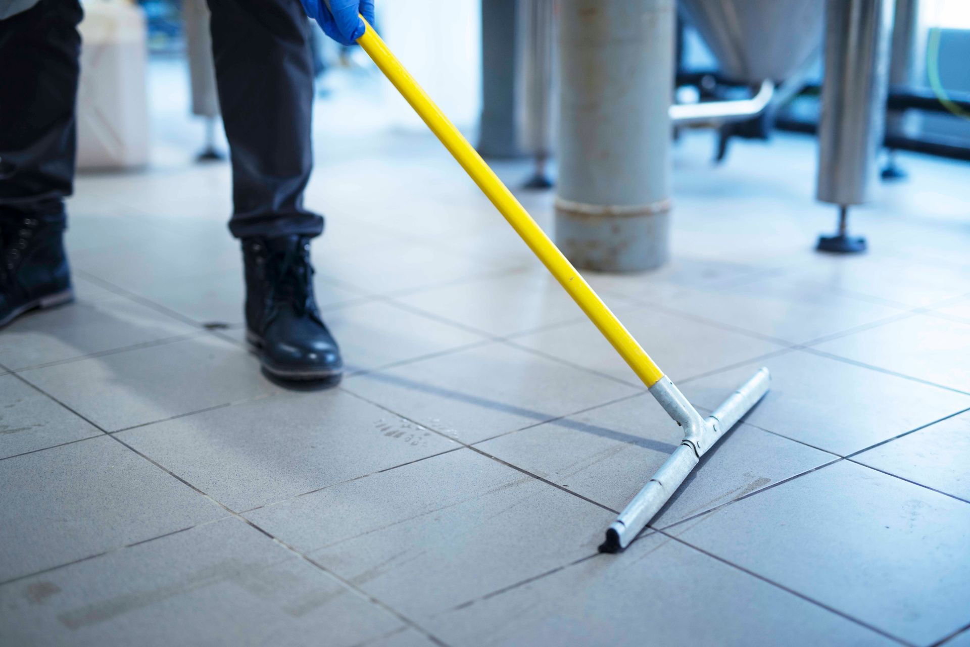 Person in blue gloves using a yellow-handled floor squeegee to clean a tiled industrial floor.