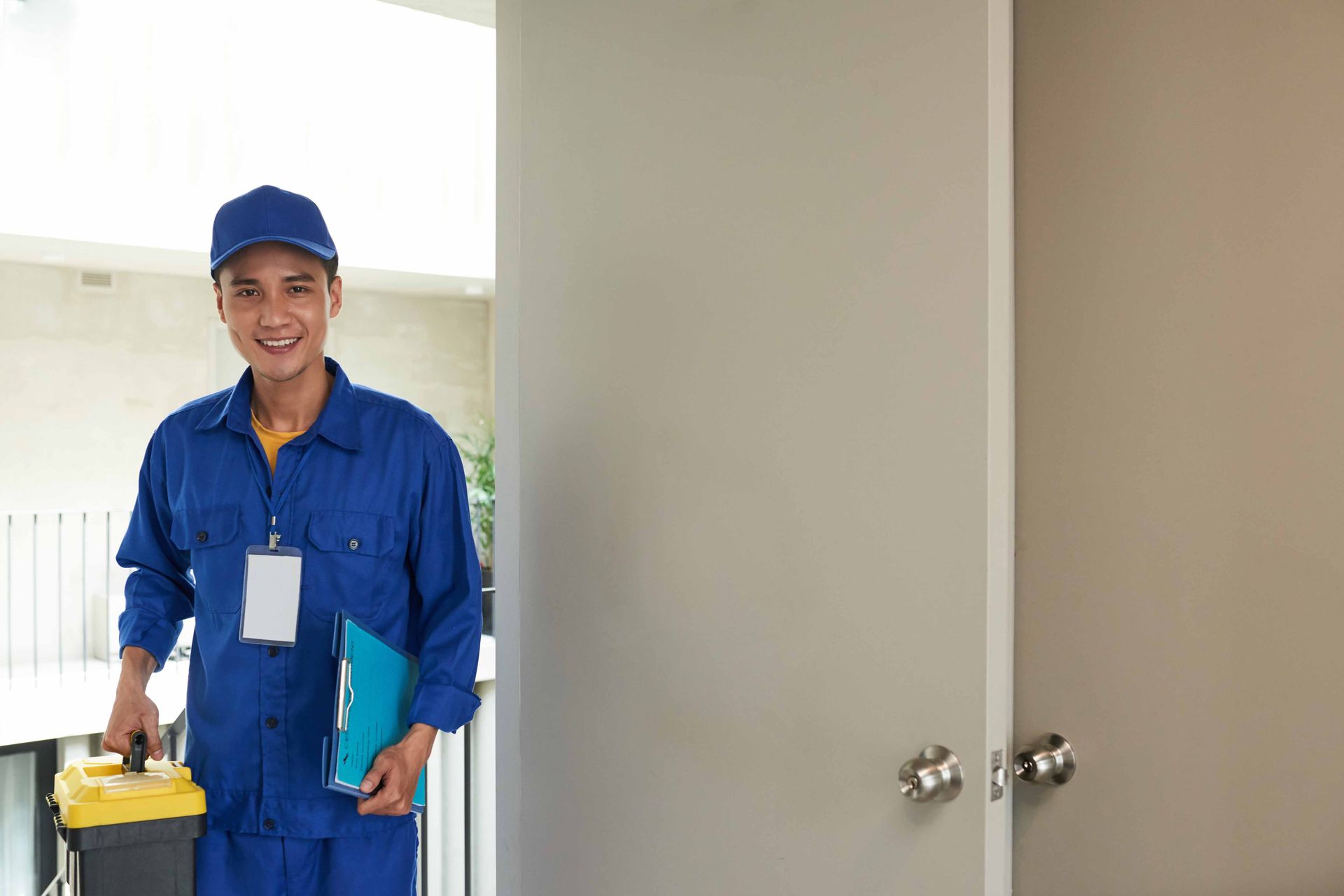Smiling person in blue work uniform with toolbox and clipboard at a door.