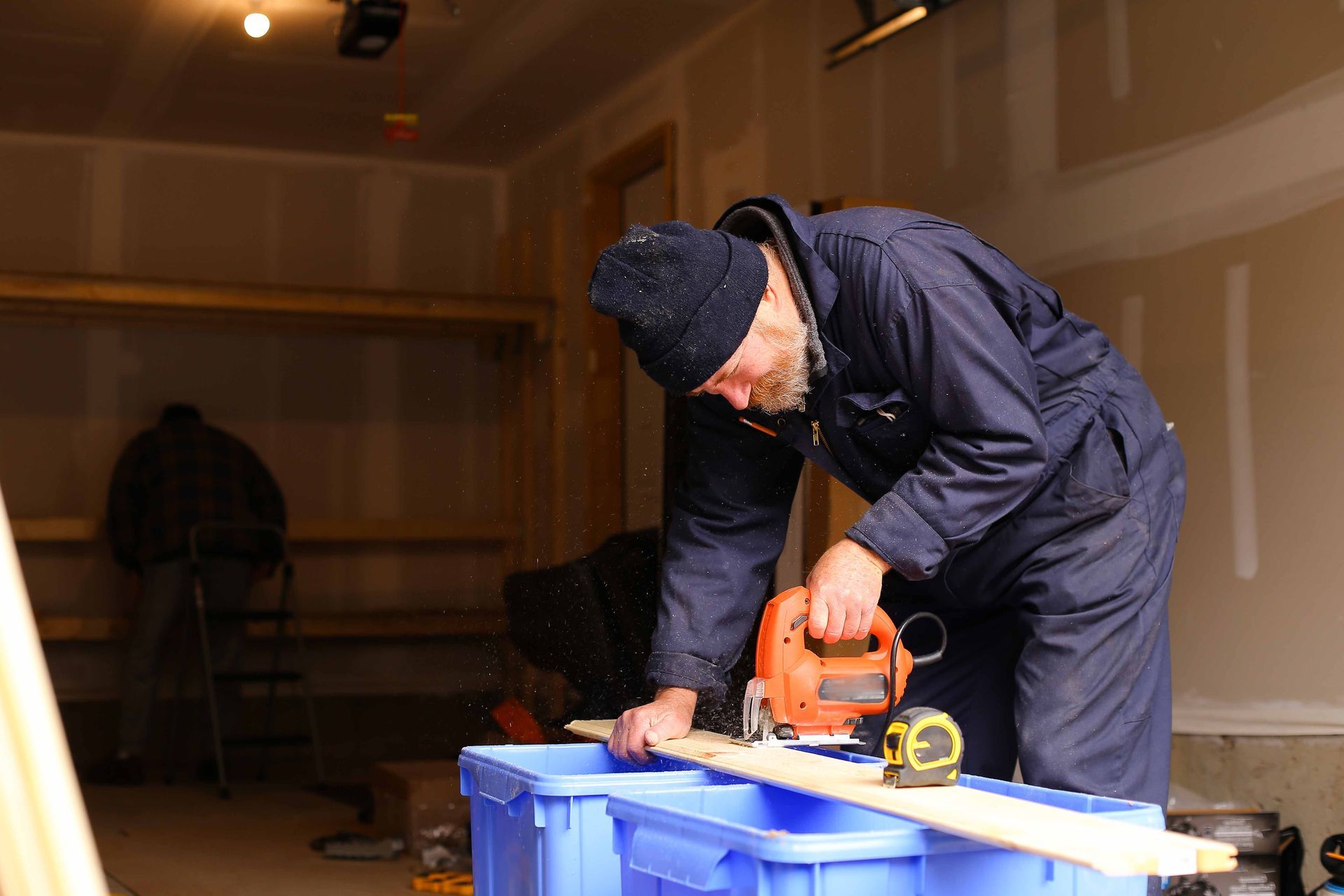 Man in blue coveralls uses a jigsaw to cut a wooden board in a garage.