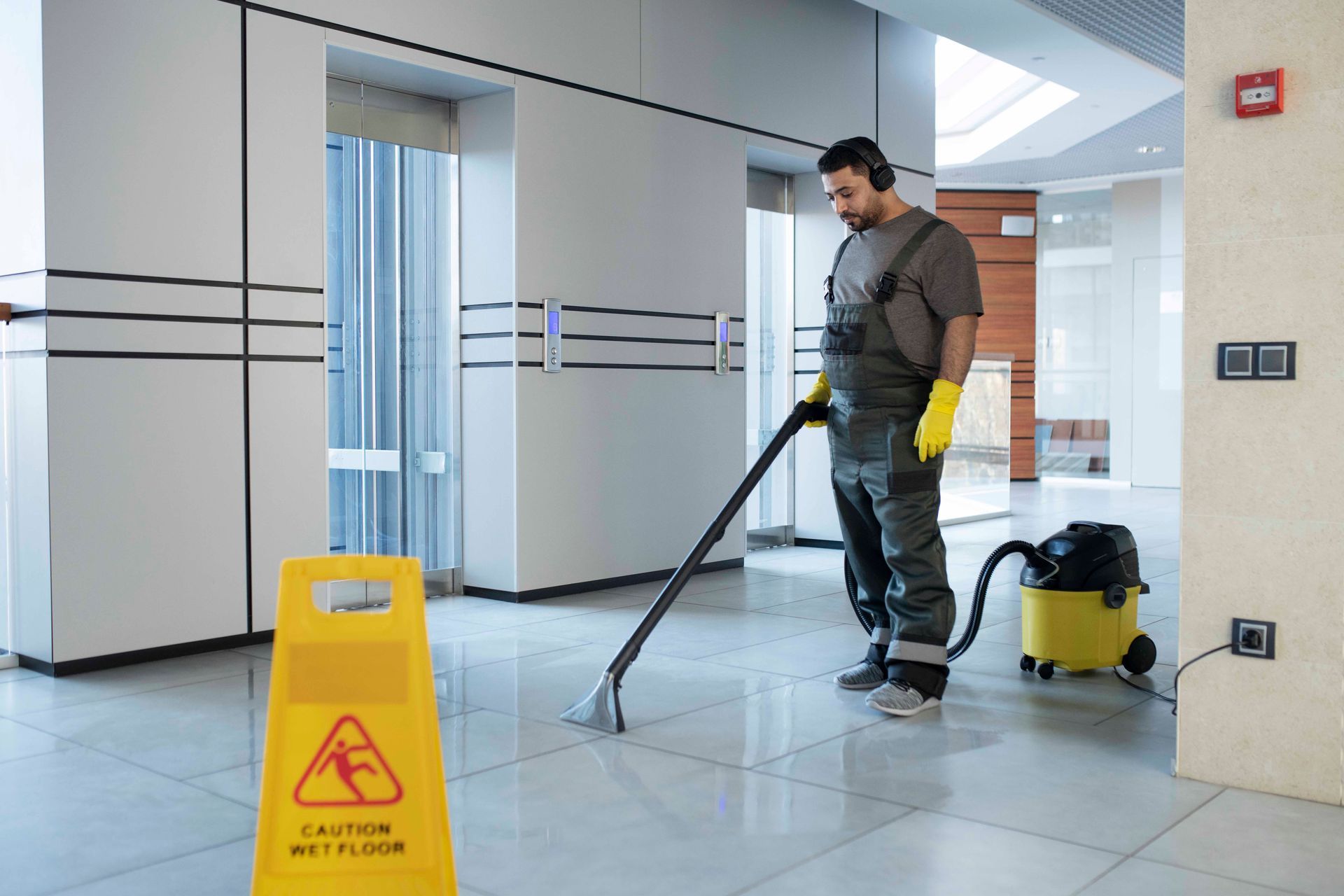 Man vacuuming wet floor in lobby, yellow caution sign nearby.