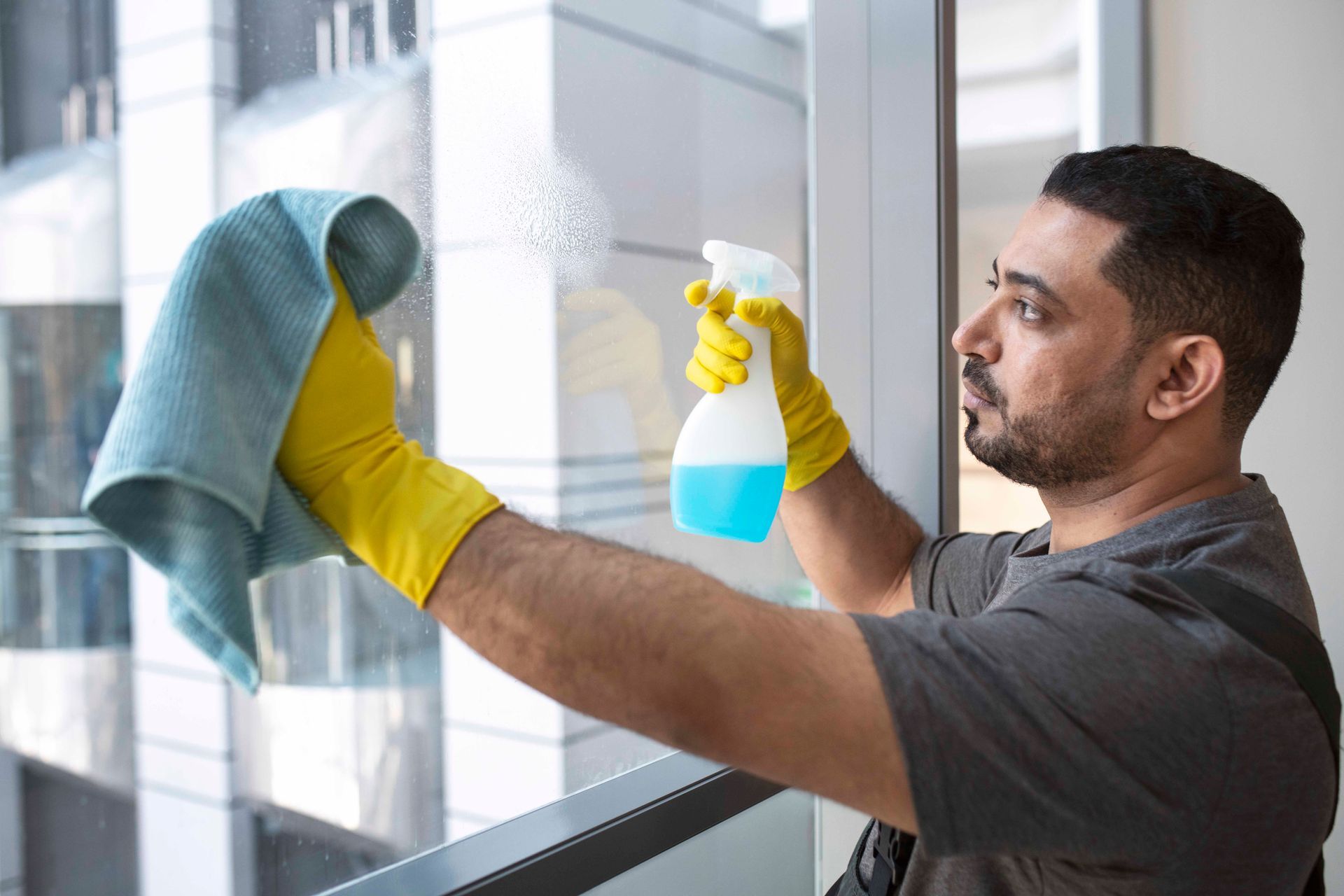 Man in yellow gloves sprays and wipes a window clean with a blue cloth.