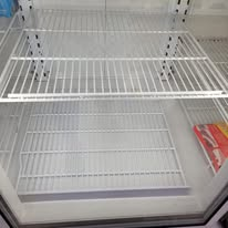 Empty refrigerator shelves. White metal shelving inside a glass-doored cooler.