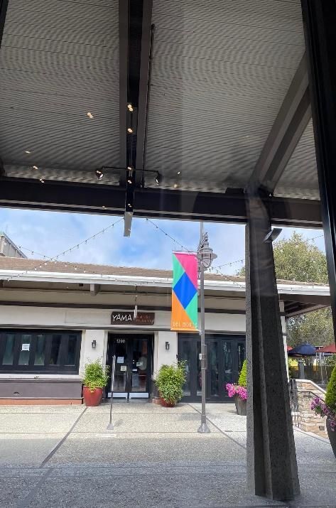 Restaurant exterior with colorful flag, potted plants, and glass windows.