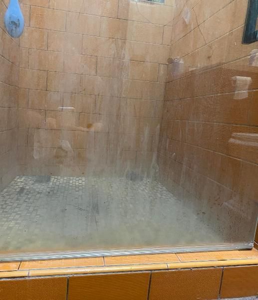 Dirty glass shower stall with light brown tile walls and floor.