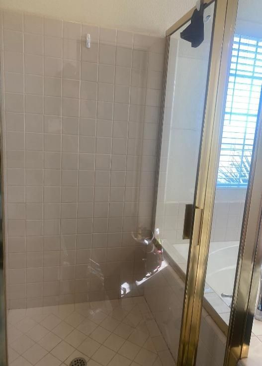 Shower stall with beige tiled walls and gold-framed glass doors next to a bathtub with a window.